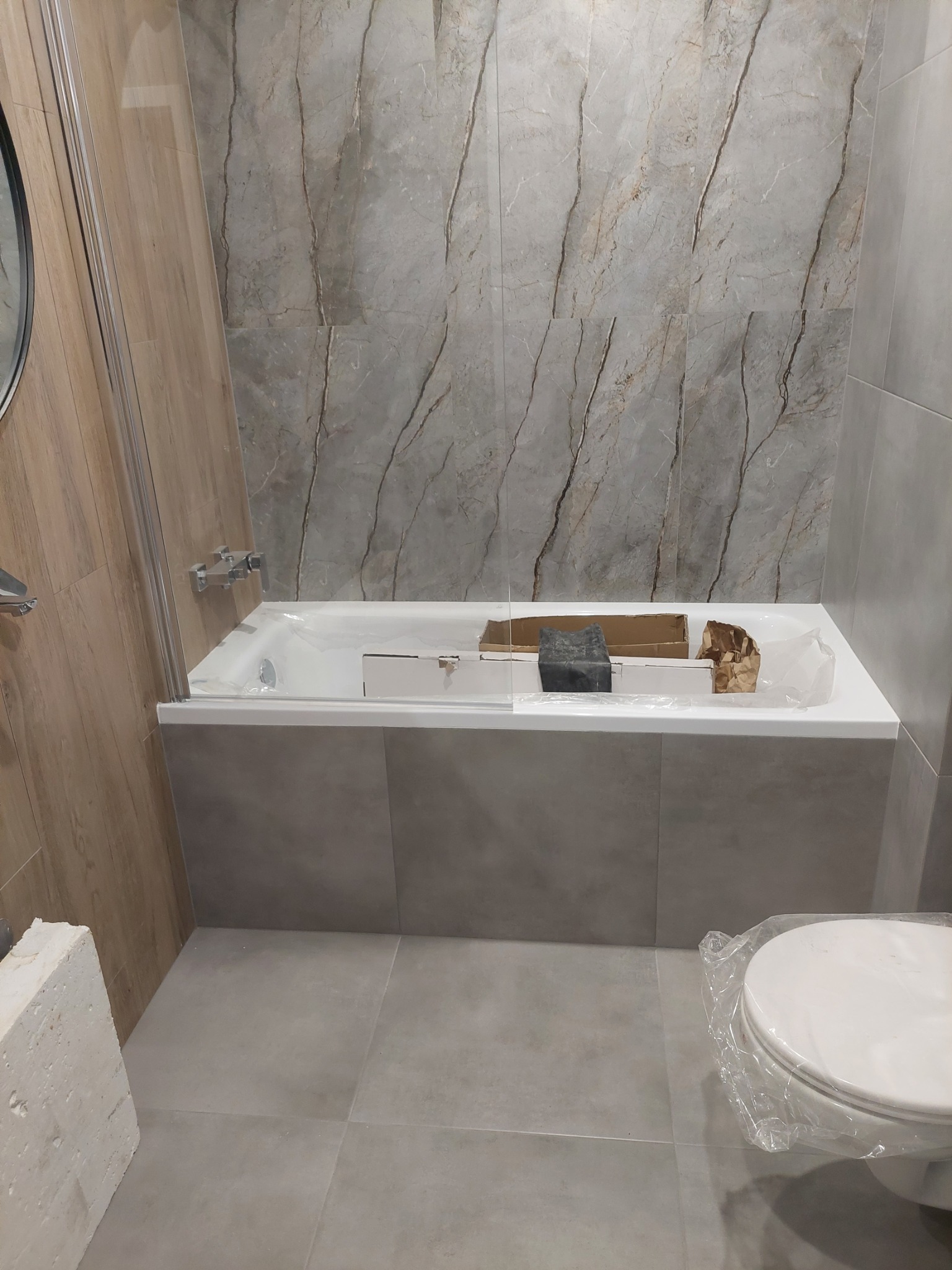 Łazienka w trakcie prac: wanna obudowana szarymi płytkami, na wannie karton, fugi i folia ochronna na toalecie, ściany wyłożone płytkami imitującymi marmur.