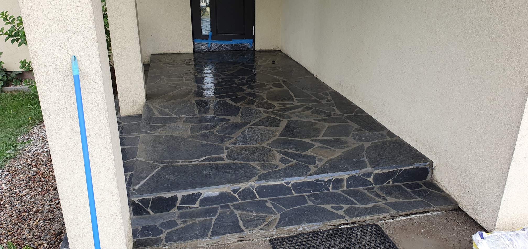 Wyczyszczony kamienny podjazd z ciemnych, nieregularnych płyt, z niebieskim mopem opartym o kolumnę, folia zabezpieczająca u progu drzwi.