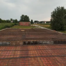 Zbrojenie stropu z widocznymi elementami dystansowymi, w tle budynek w trakcie budowy i krajobraz wiejski pod pochmurnym niebem.