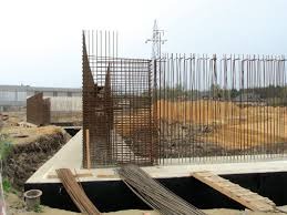 Zbrojenie fundamentów budynku w trakcie budowy, widoczne pręty stalowe i szalunki, w tle słup wysokiego napięcia.