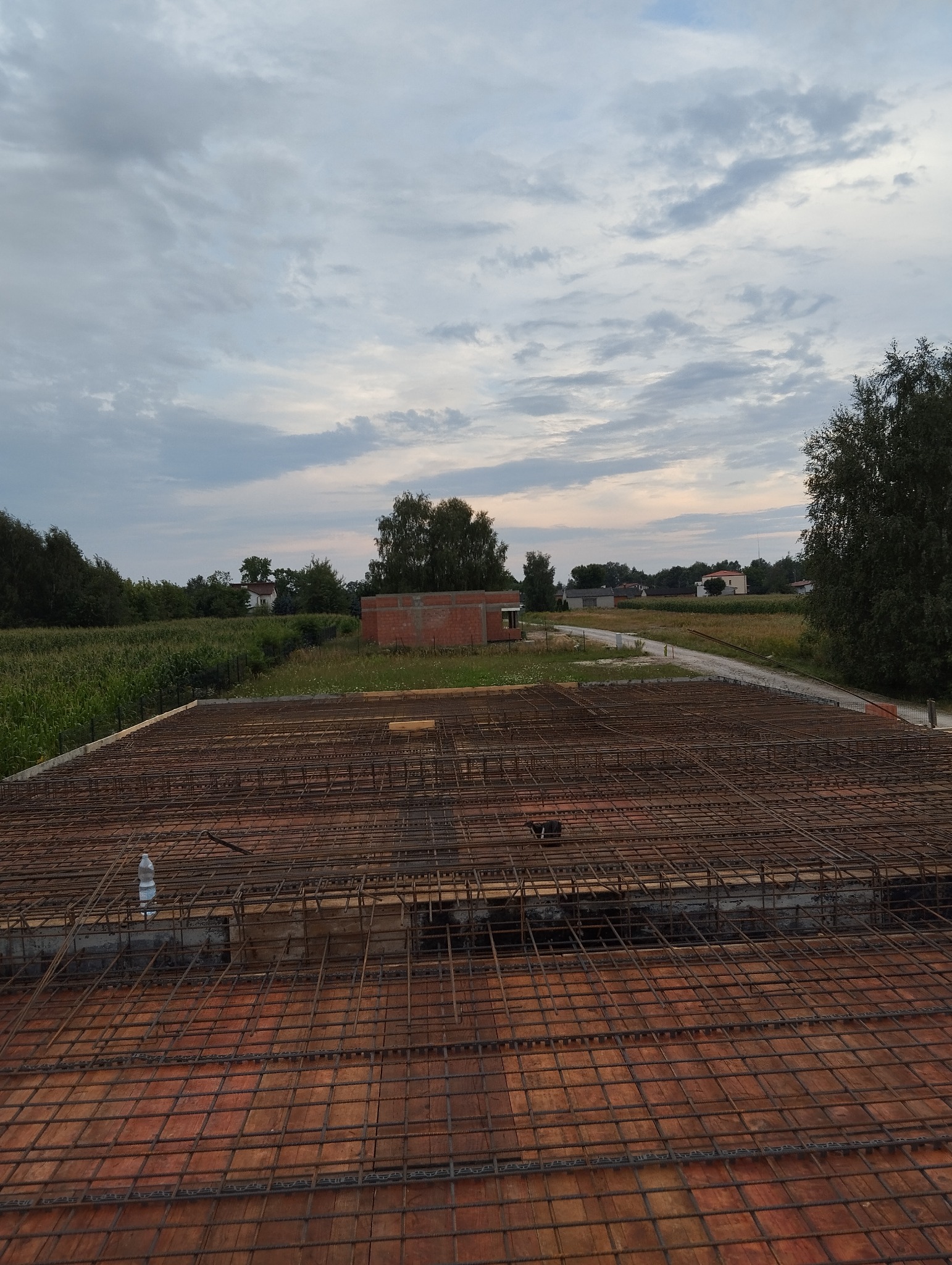 Siatka zbrojeniowa ułożona na szalunku z desek, widoczna butelka wody, w tle niedokończony budynek z czerwonej cegły i zielone pola pod zachmurzonym niebem.
