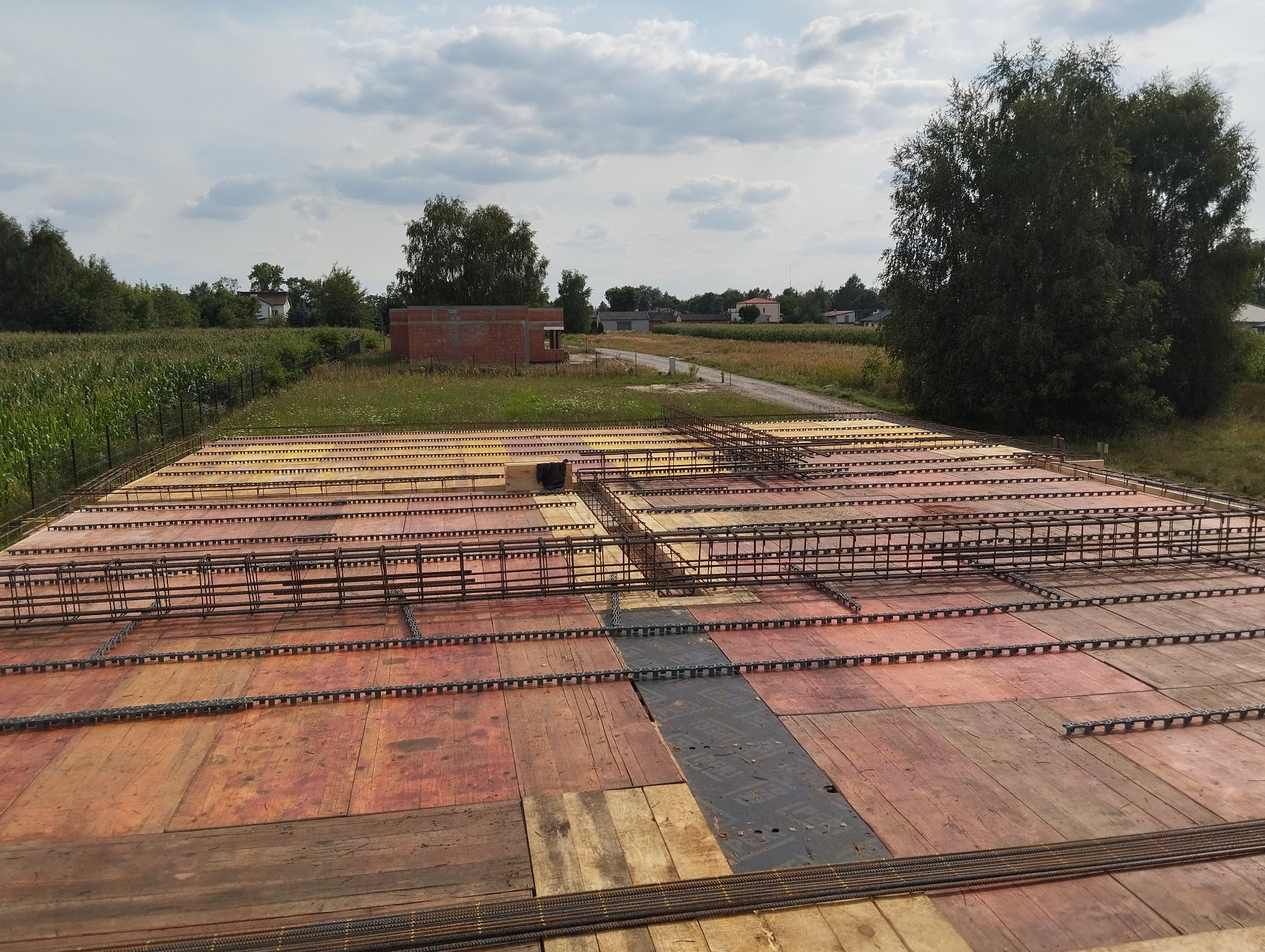 Przygotowanie szalunku i zbrojenia płyty fundamentowej na budowie domu jednorodzinnego, widok z góry na drewniane płyty i ułożone pręty zbrojeniowe z dystansami, w tle budynek w stanie surowym...