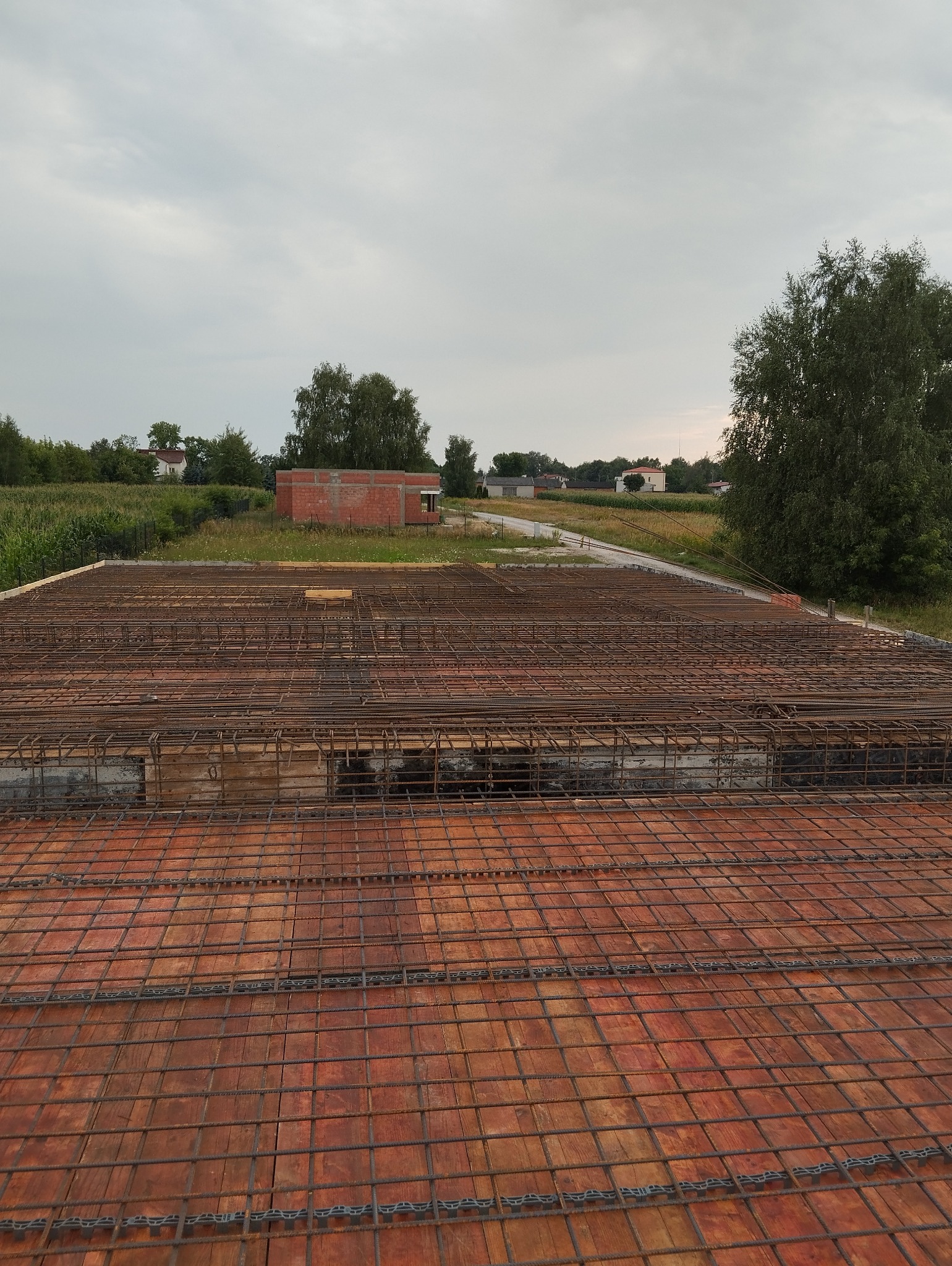 Zbrojenie stropu z widocznymi elementami dystansowymi, w tle budynek w trakcie budowy i krajobraz wiejski pod pochmurnym niebem.