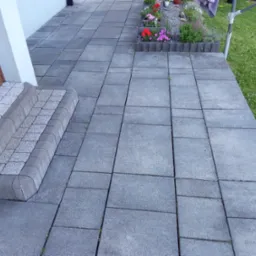 Szara kostka brukowa ułożona na podjeździe z widocznymi schodami wejściowymi i rabatą kwiatową w tle.