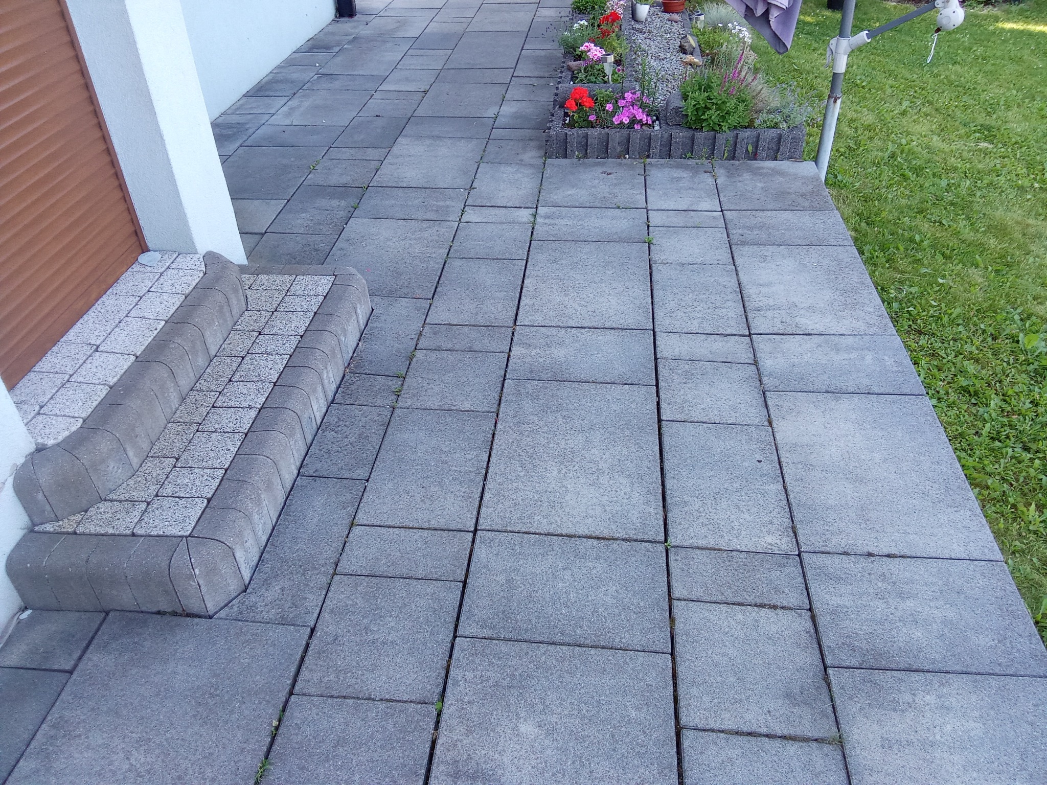 Szara kostka brukowa ułożona na podjeździe z widocznymi schodami wejściowymi i rabatą kwiatową w tle.