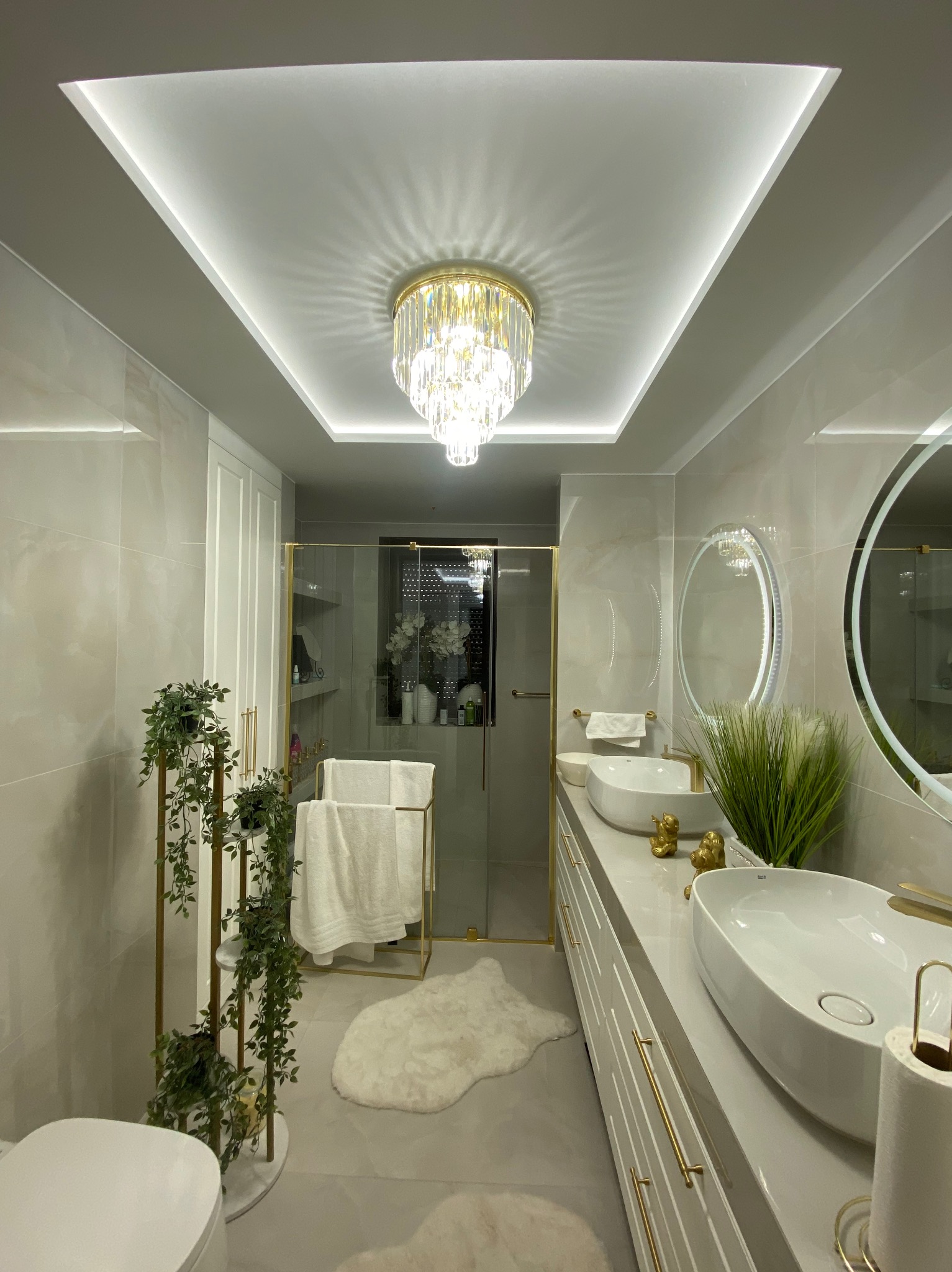 Nowoczesna łazienka z podwójną umywalką nablatową, oświetleniem LED w suficie podwieszanym i kryształowym żyrandolem, wykończona w jasnych szarościach i bieli z akcentami złota.