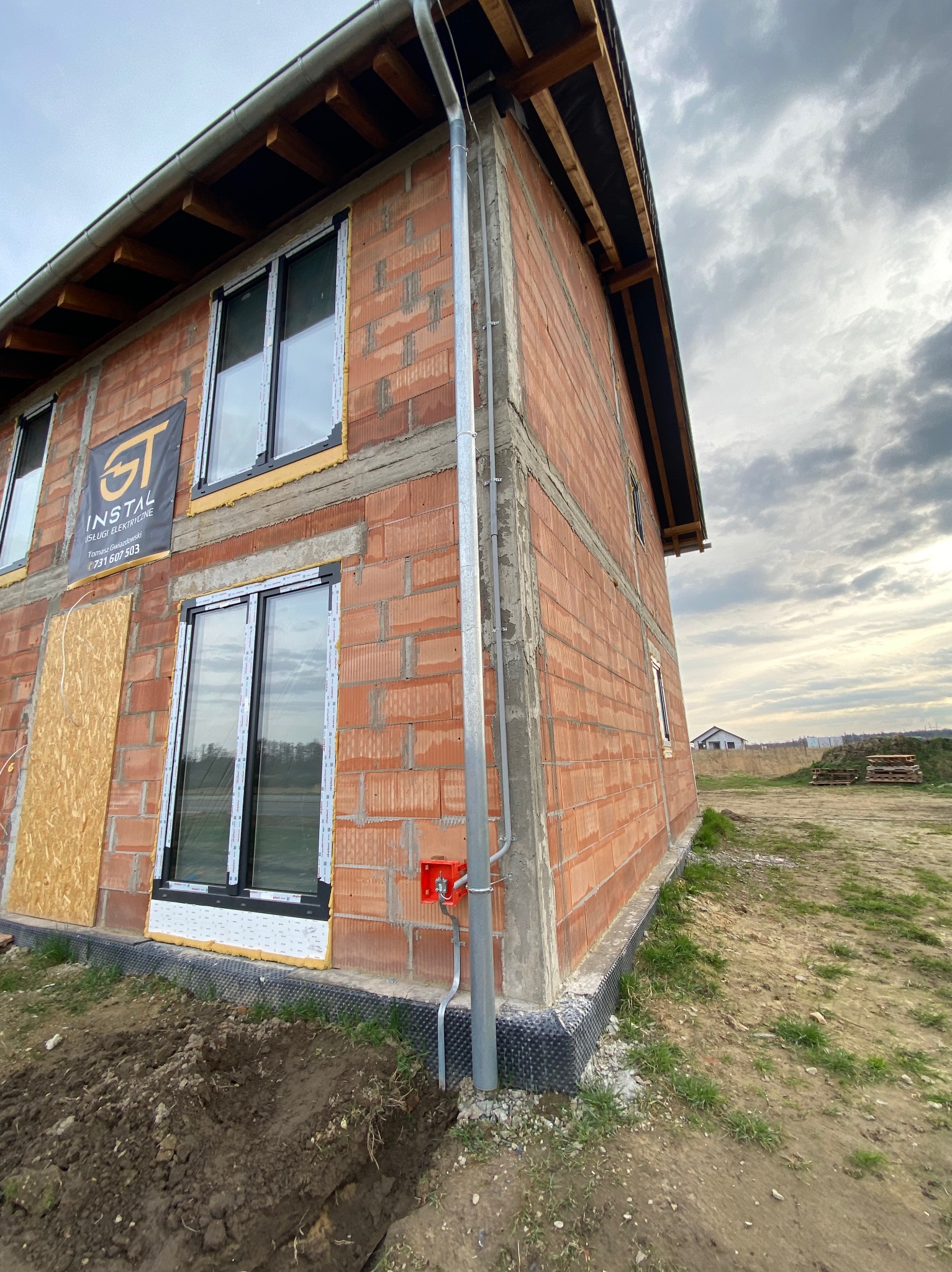 Surowy budynek z czerwonej cegły w trakcie budowy, widoczna instalacja odgromowa z pomarańczową puszką przyłączeniową, okna z czarnymi ramami, logo firmy na ścianie i wykop w ziemi.