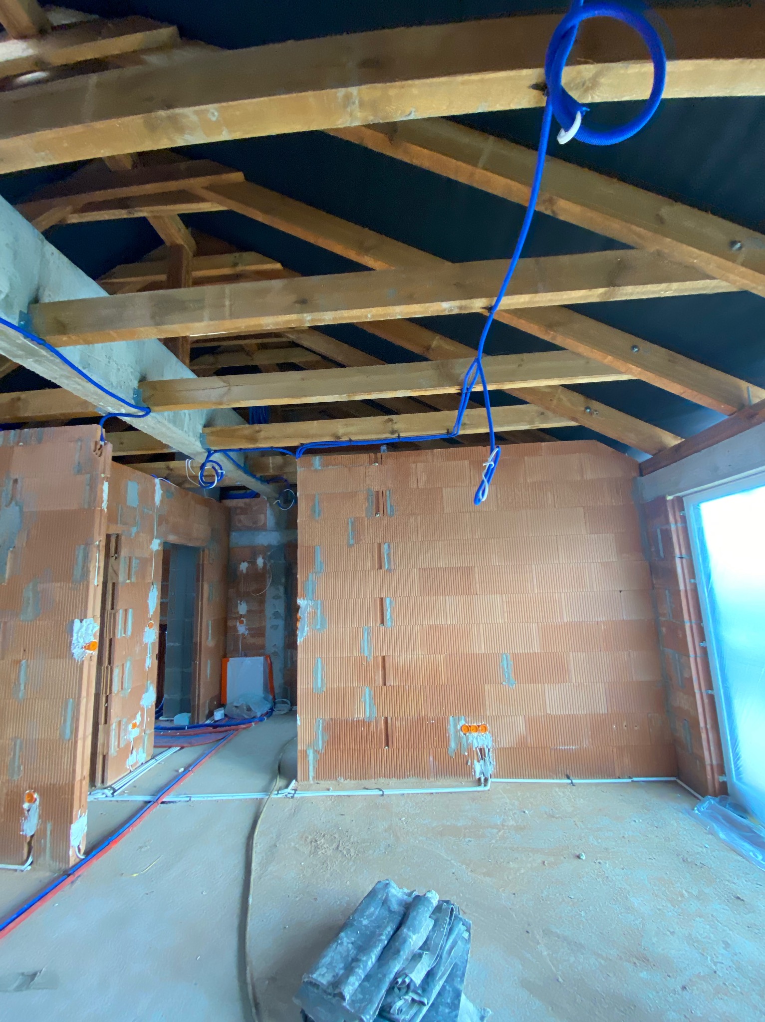 Wnętrze w trakcie instalacji elektrycznej i wodnej: widoczne ceglane ściany, drewniana konstrukcja dachu z czarną membraną, niebieskie rury instalacji wodnej i białe rury instalacji elektrycznej...