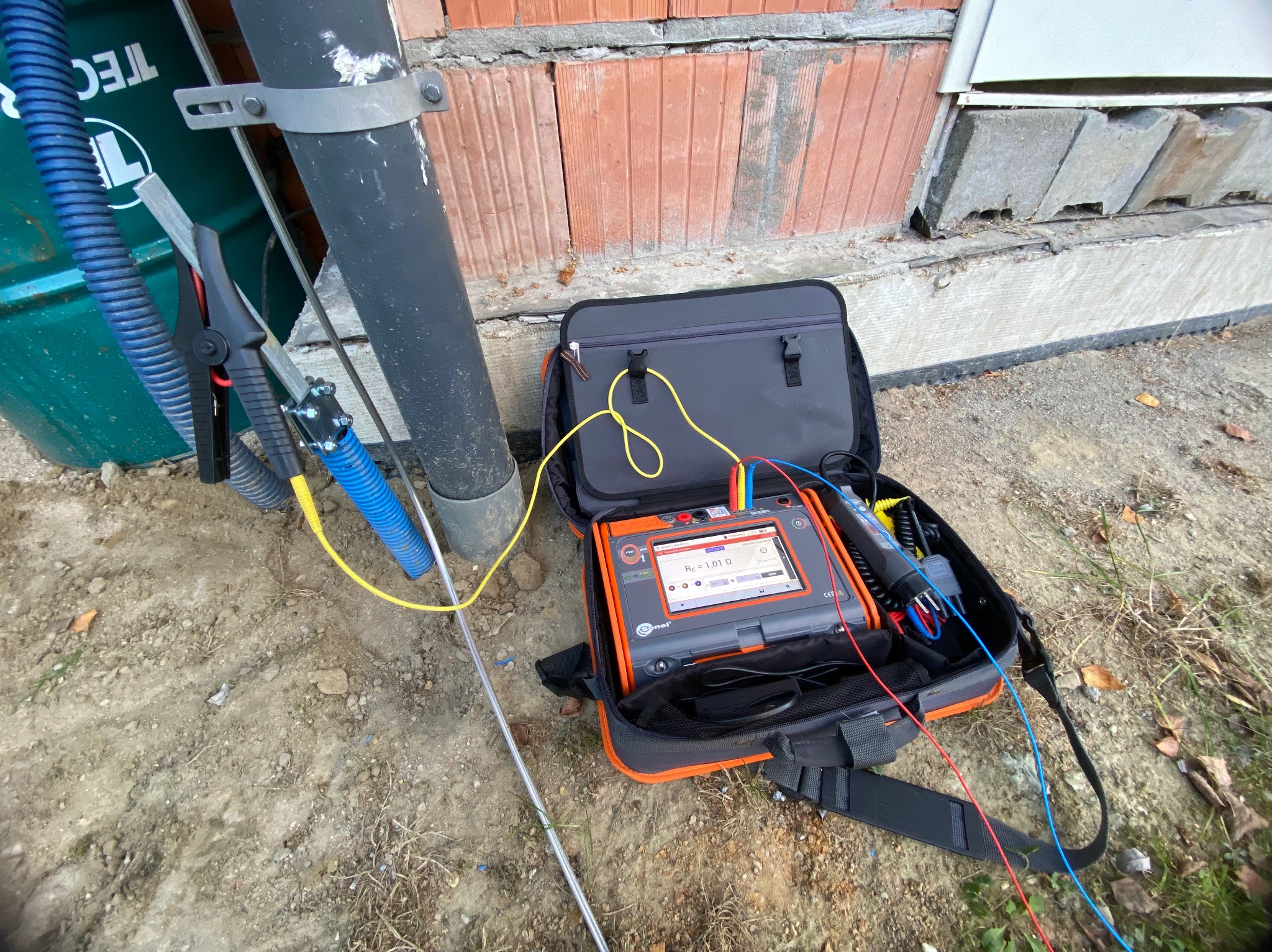 Miernik rezystancji uziemienia Sonel w torbie, podłączony przewodami do uziomu przy ścianie budynku z cegły kratówki.
