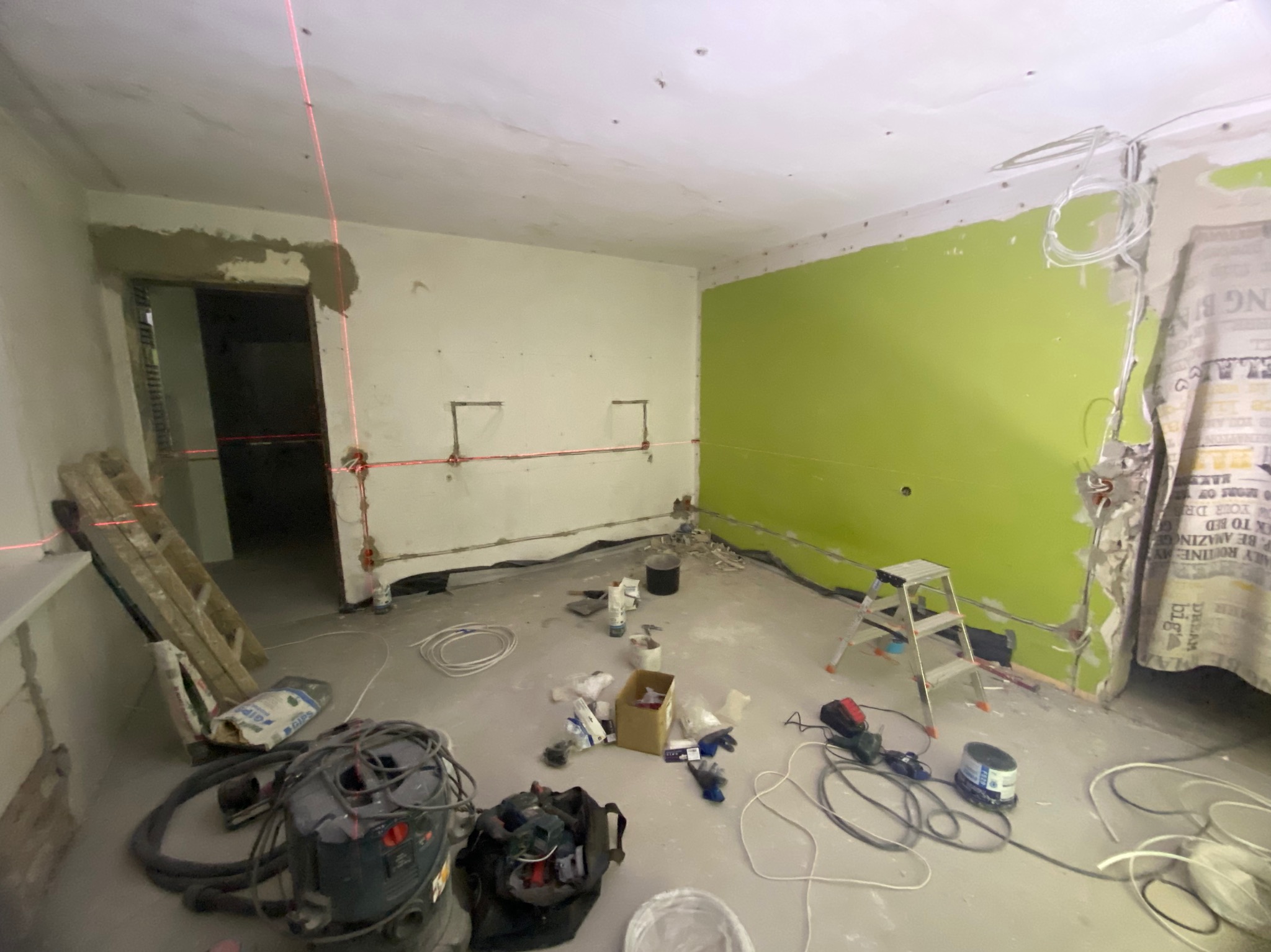 Wnętrze w trakcie remontu z widocznymi przewodami elektrycznymi, poziomica laserowa wyznacza linię, drabina oparta o ścianę, na podłodze rozłożone narzędzia i materiały budowlane, ściany częściowo...