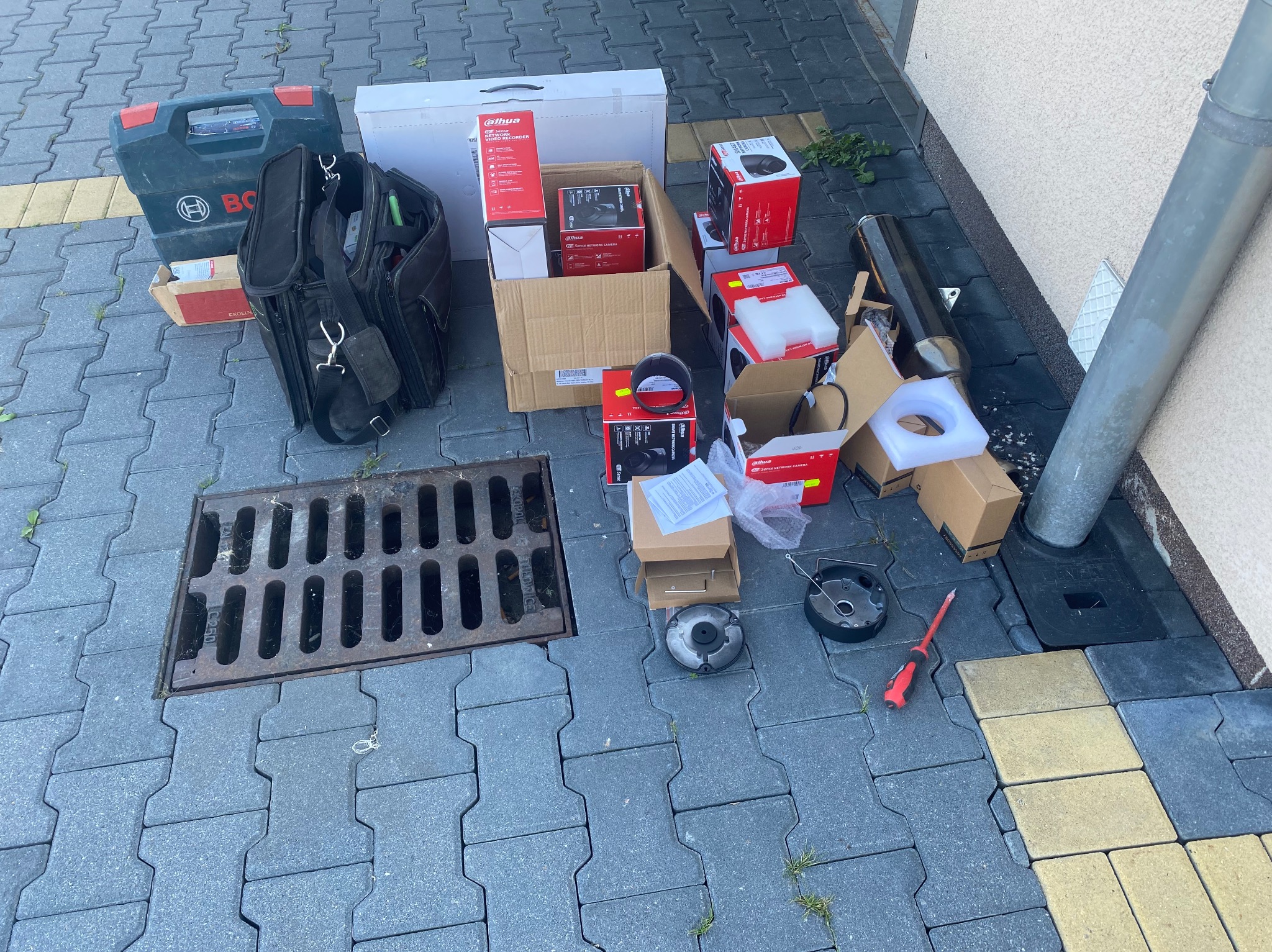 Rozpakowane kamery monitoringu Dahua, rejestrator, narzędzia i torba monterska leżące na kostce brukowej obok studzienki kanalizacyjnej