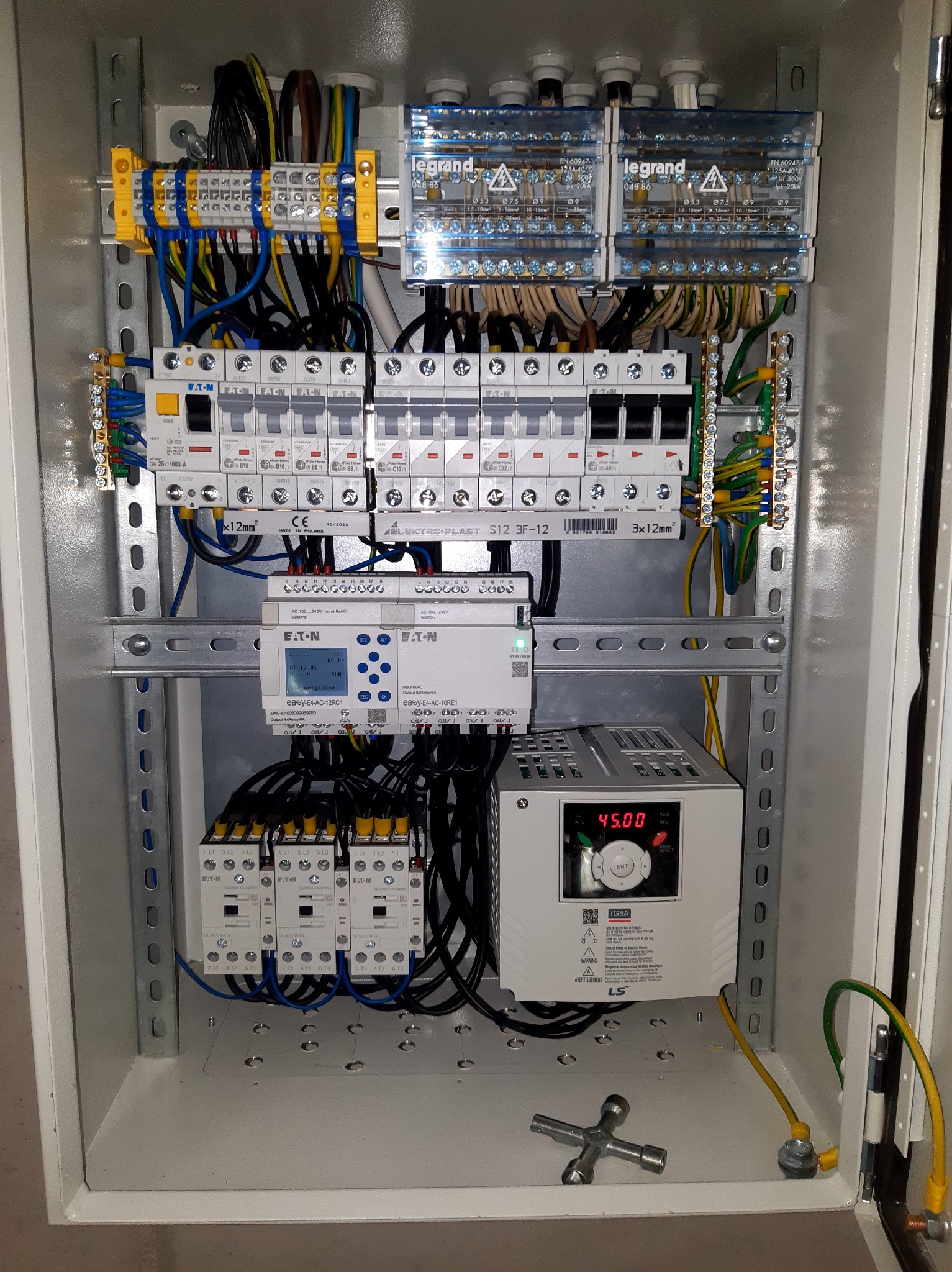 Rozdzielnica elektryczna w metalowej obudowie, widoczne bezpieczniki, przekaźniki, sterownik PLC Eaton easyE4, falownik LS iG5A z wyświetlaczem wskazującym 45.00, przewody w różnych kolorach...