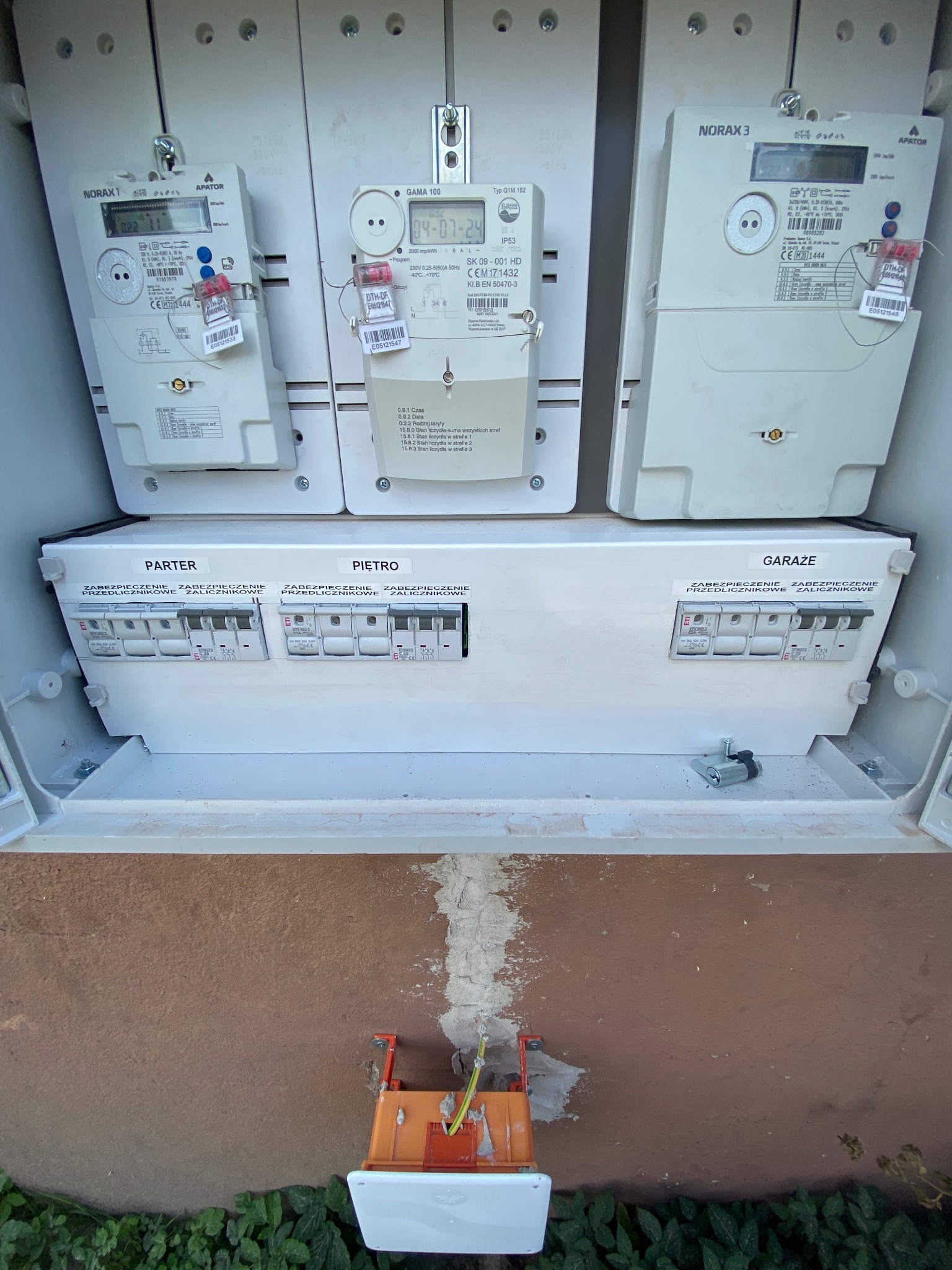 Skrzynka z trzema licznikami energii elektrycznej NORAX i GAMA, oraz zabezpieczeniami obwodów parteru, piętra i garażu, zamontowana na elewacji budynku, z widocznym przewodem zasilającym w dolnej...