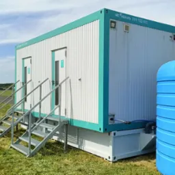 Przenośna toaleta kontenerowa z turkusowymi obramowaniami i metalowymi schodami, na trawiastym terenie z widocznym samolotem na pasie startowym w tle.