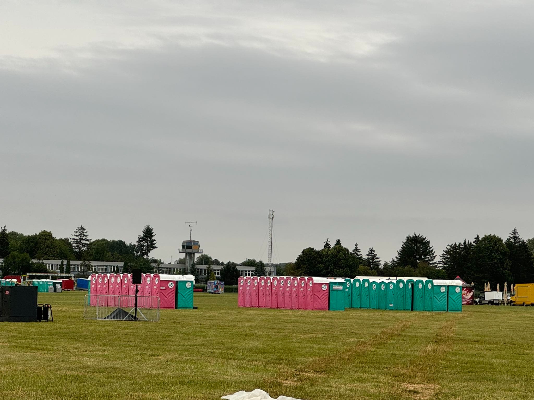 Ustawione rzędy przenośnych toalet w kolorach różowym i zielonym na trawiastym terenie, w tle budynek i drzewa pod pochmurnym niebem, prawdopodobnie na terenie imprezy plenerowej.