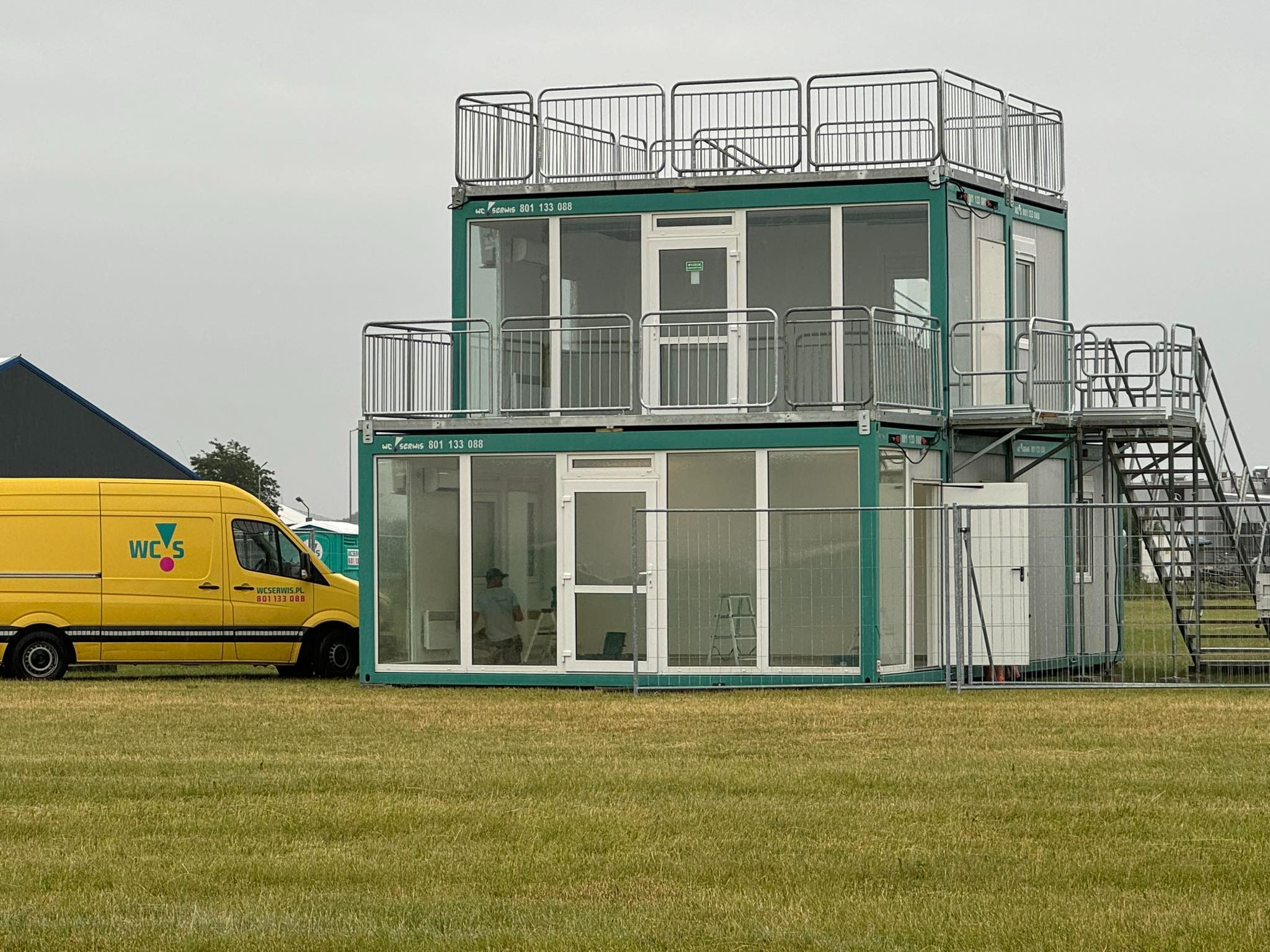 Dwukondygnacyjny, zielony kontener sanitarny z balkonami i schodami na zewnątrz, widoczny na trawiastym terenie, z żółtym samochodem serwisowym w tle.