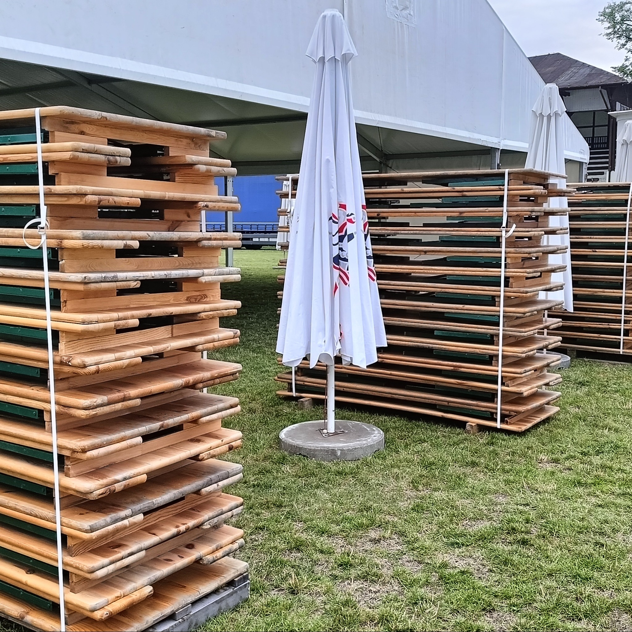 Stosy złożonych, drewnianych stołów cateringowych z zielonymi elementami konstrukcyjnymi, zabezpieczone białymi pasami, obok białego parasola na trawie, z białym namiotem w tle.