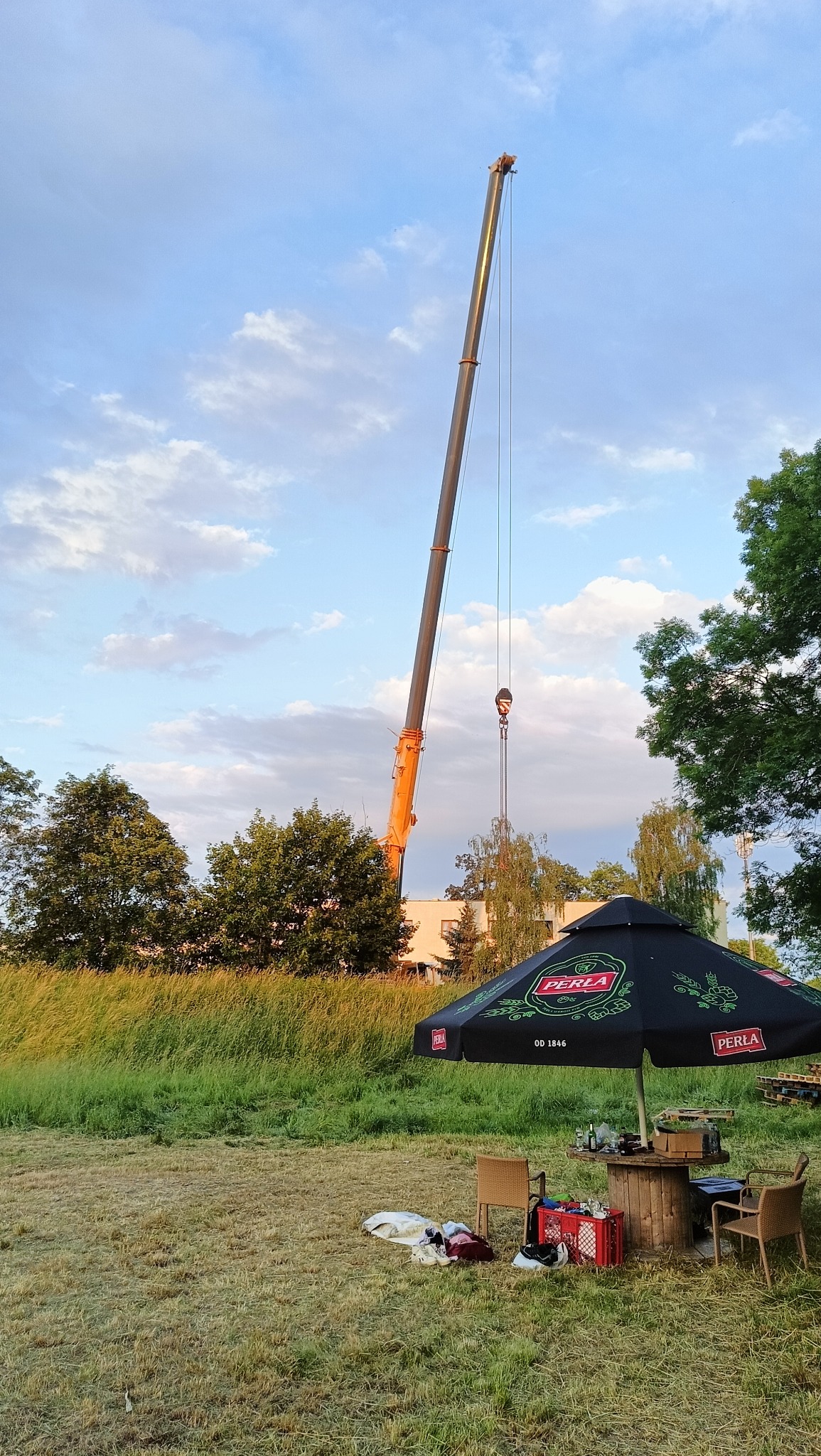 Żuraw budowlany na tle letniego krajobrazu z parasolem Perła i stolikiem z krzesłami.