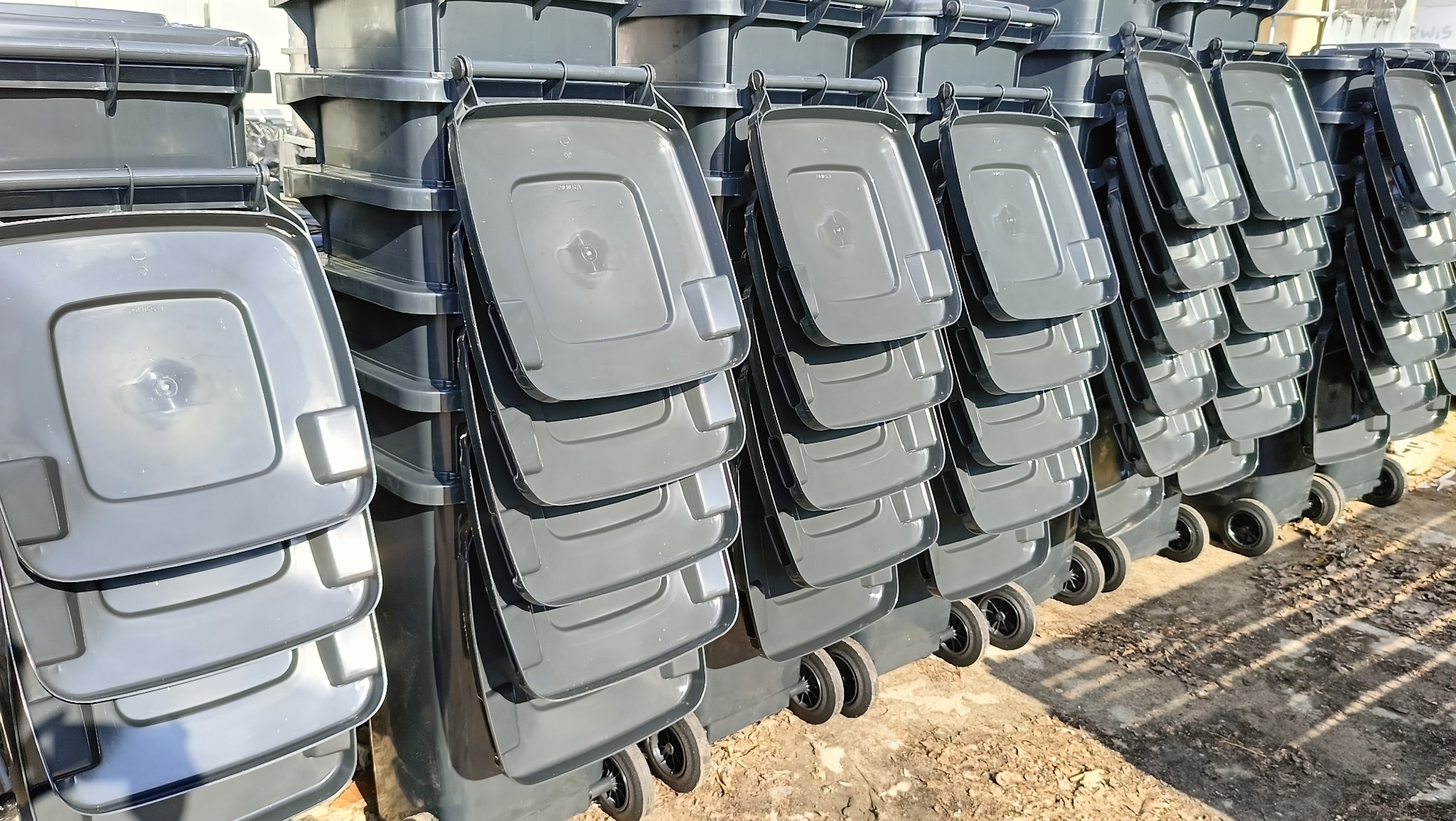 Rząd szarych pojemników na śmieci z pokrywami i kółkami, ustawionych jeden za drugim, w magazynie lub na placu budowy.