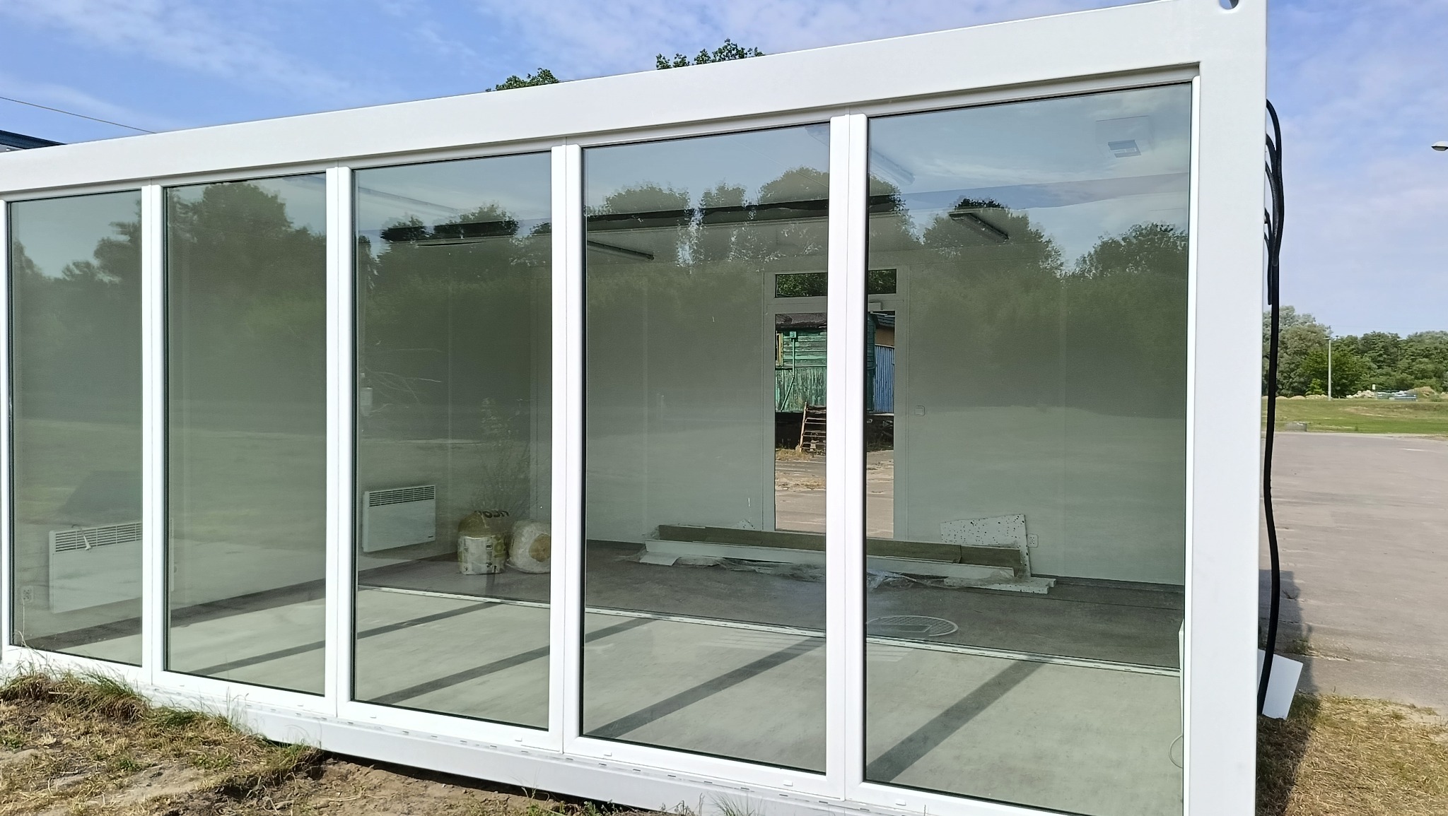 Biały pawilon kontenerowy z dużymi oknami, widoczne grzejniki elektryczne wewnątrz, teren z zielenią w tle.