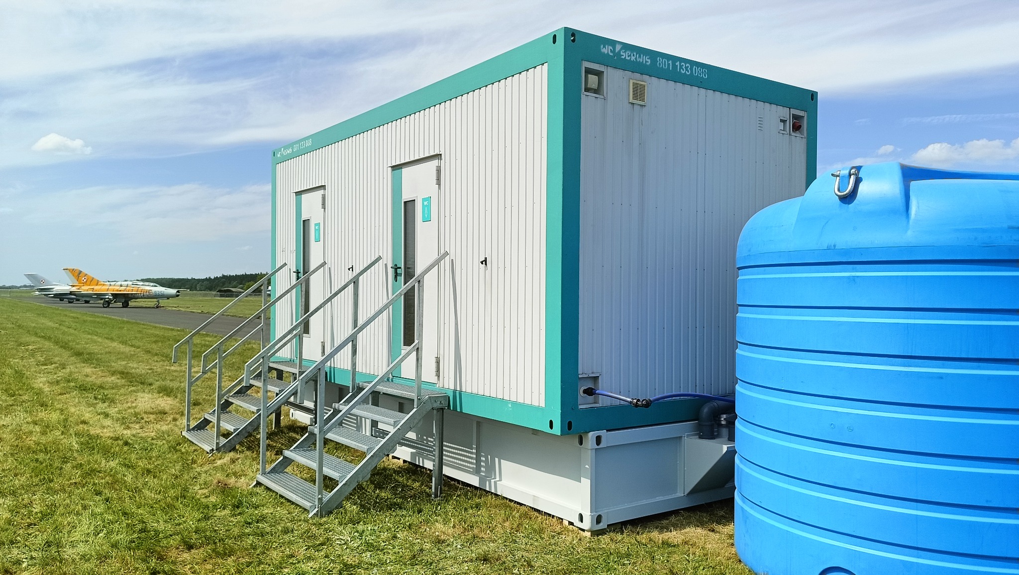 Przenośna toaleta kontenerowa z turkusowymi obramowaniami i metalowymi schodami, na trawiastym terenie z widocznym samolotem na pasie startowym w tle.