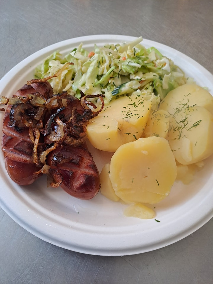 Porcja obiadowa na papierowym talerzu: grillowana kiełbasa z karmelizowaną cebulą, gotowane ziemniaki posypane koperkiem oraz surówka z białej kapusty.