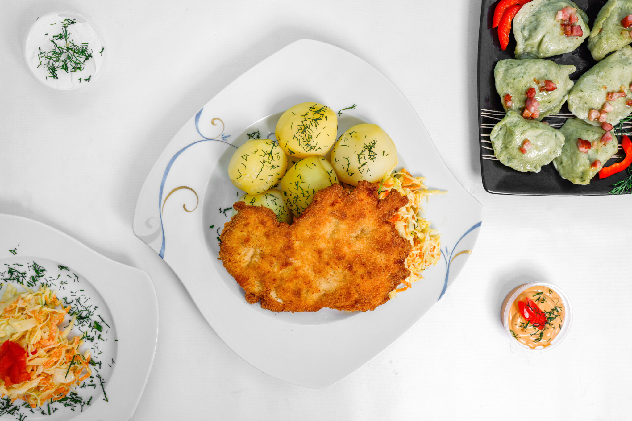 Widok z góry na talerz z panierowanym kotletem, gotowanymi ziemniakami posypanymi koperkiem, surówką z kapusty, obok pierogi ze szpinakiem z podsmażonym boczkiem i czerwoną papryką oraz sosy...