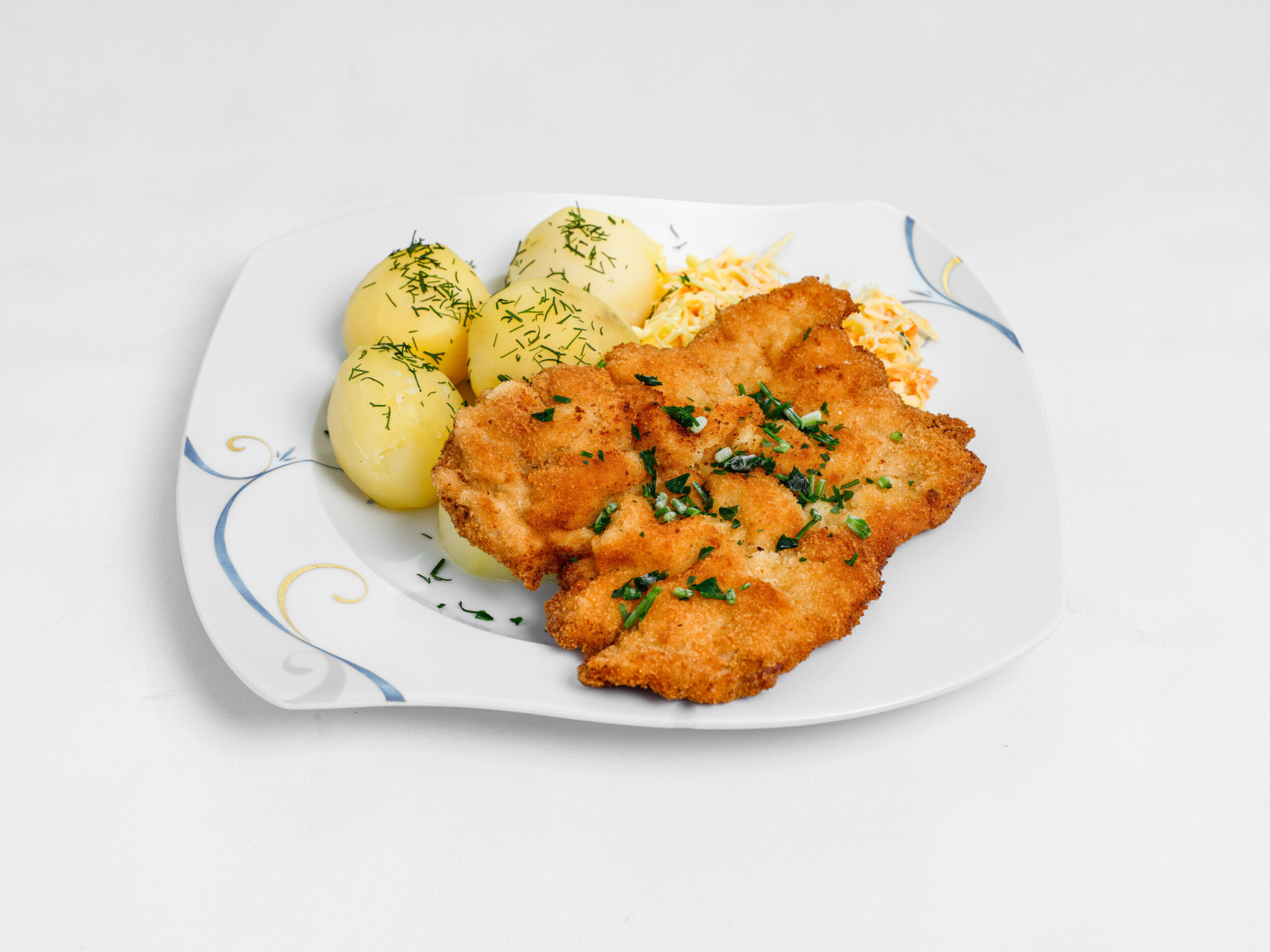 Dwa kotlety schabowe posypane świeżym szczypiorkiem, podane z gotowanymi ziemniakami posypanymi koperkiem i surówką z kapusty na białym talerzu z niebiesko-złotym wzorem.