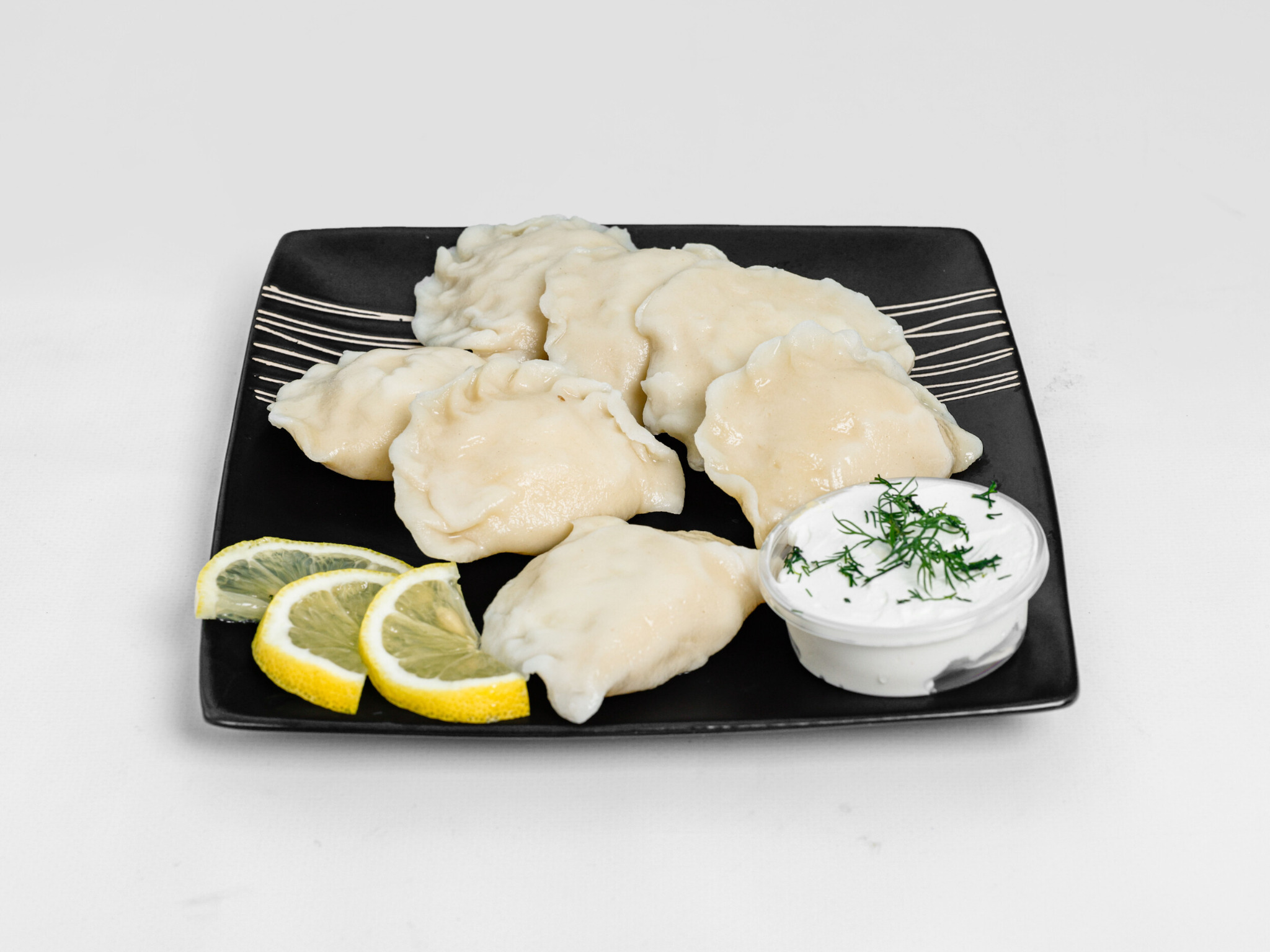 Kluski na parze, podane na czarnym talerzu z sosem śmietanowym posypanym koperkiem i ćwiartkami cytryny.