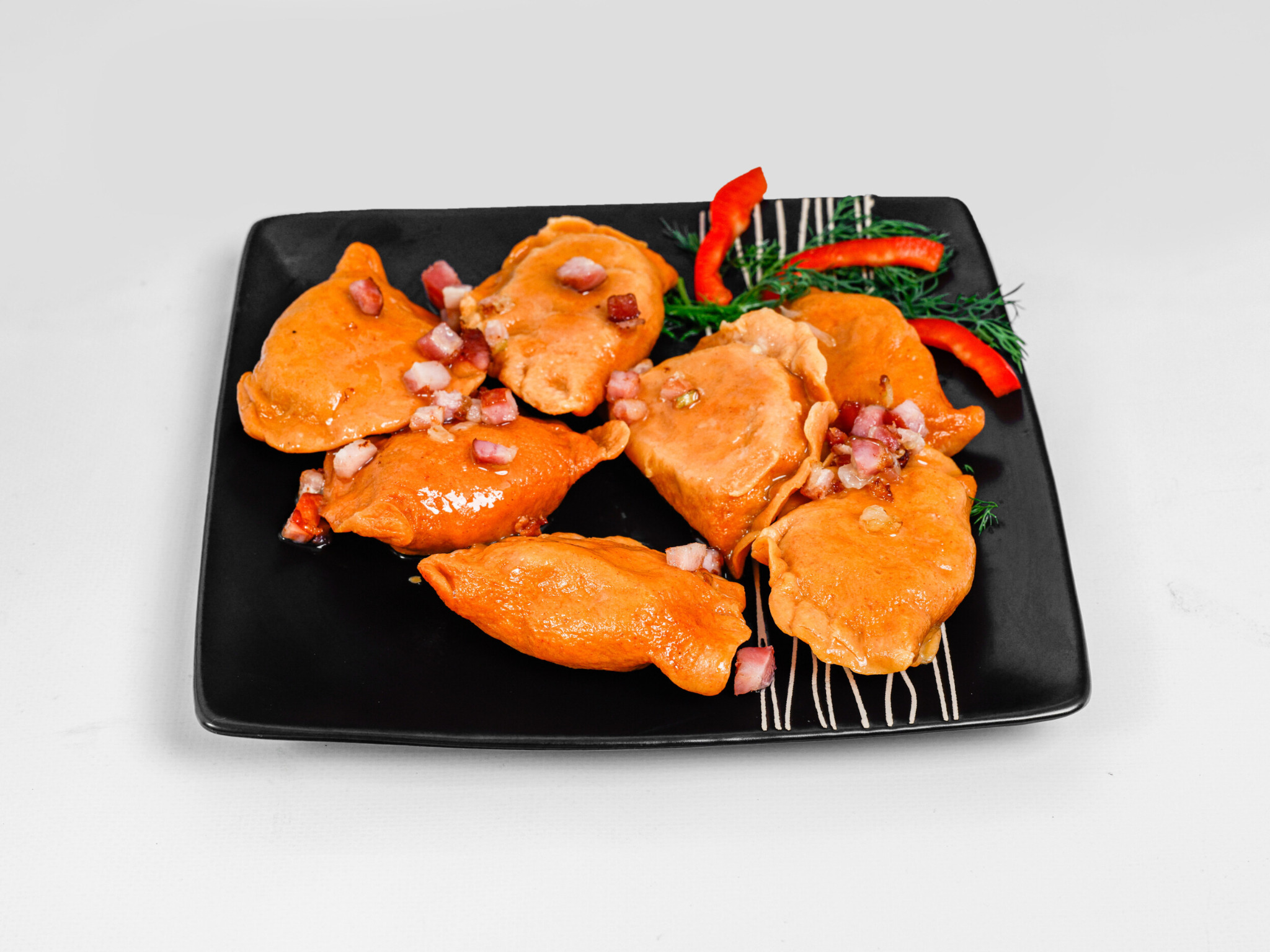 Apetyczne pierogi z mięsem i skwarkami na czarnym, kwadratowym talerzu, udekorowane czerwoną papryką i koperkiem, na białym tle.