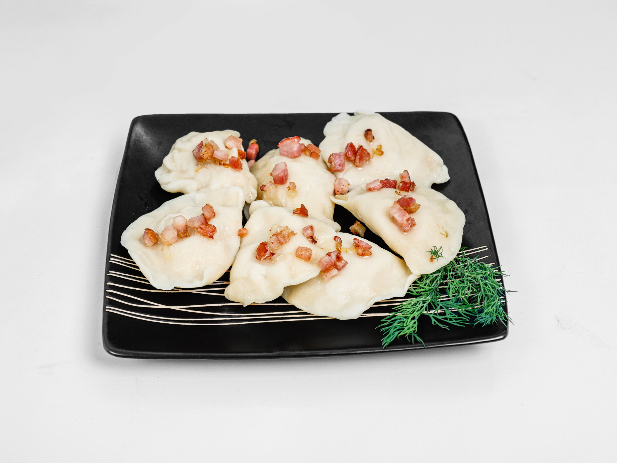Świeżo ugotowane pierogi okraszone skwarkami na czarnym, kwadratowym talerzu z dodatkiem gałązki świeżego kopru.