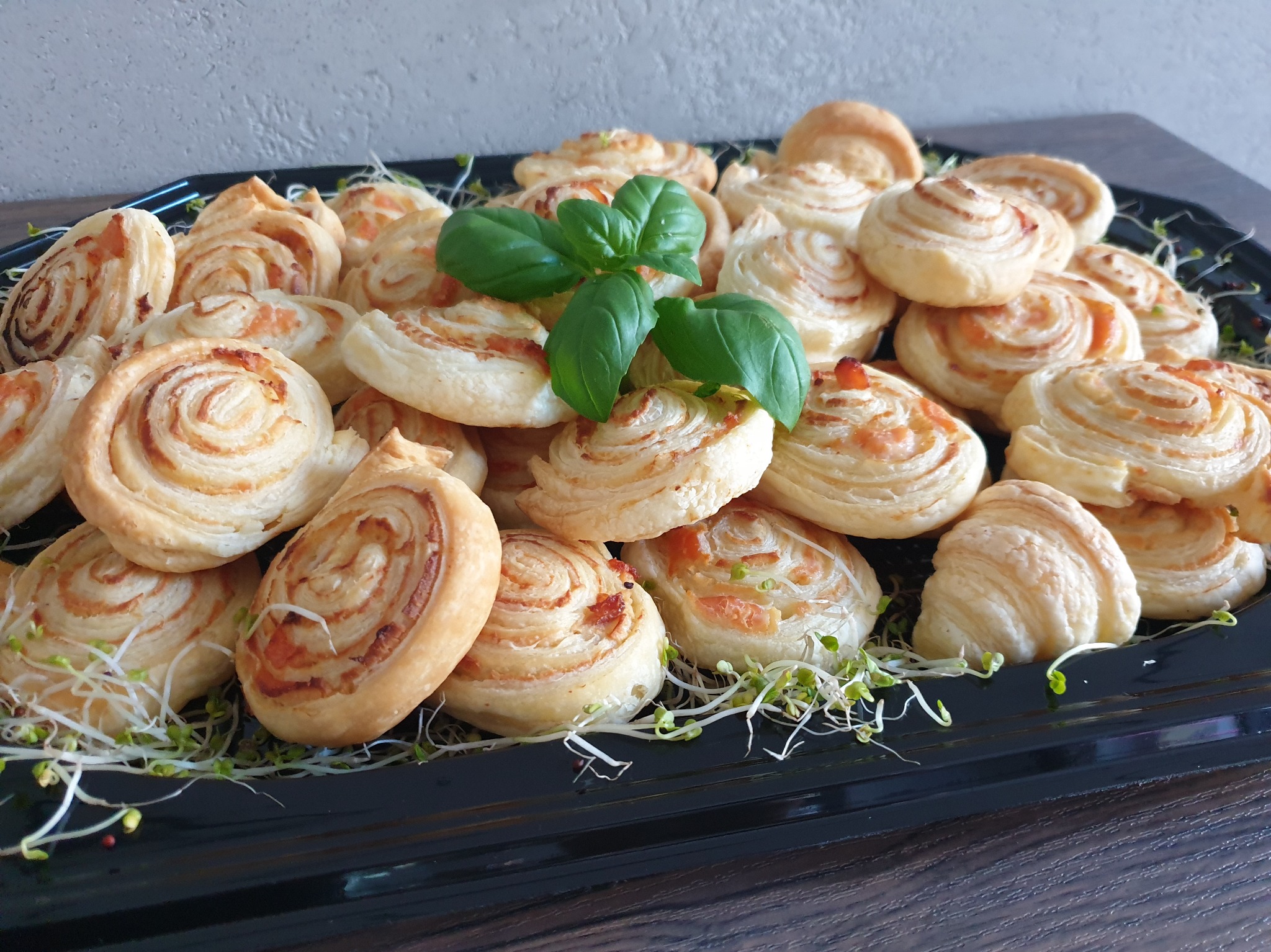 Apetyczne roladki z ciasta francuskiego z farszem, ułożone na czarnej tacy z dekoracją ze świeżych kiełków i listków bazylii.