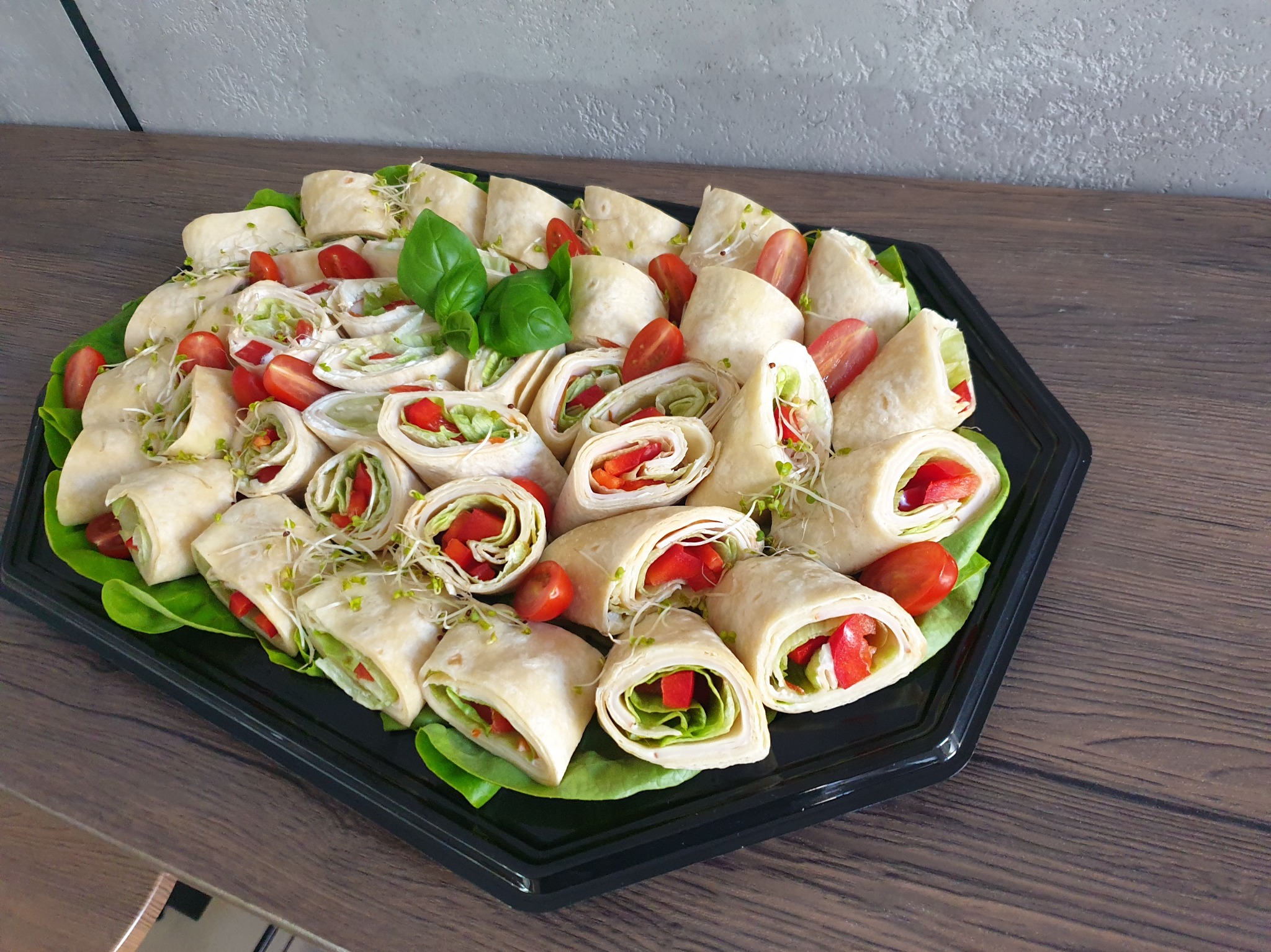 Elegancko ułożone roladki z tortilli z warzywami (papryka, sałata, kiełki) i pomidorkami koktajlowymi na czarnej tacy, udekorowane liśćmi sałaty i bazylii, na drewnianym blacie.