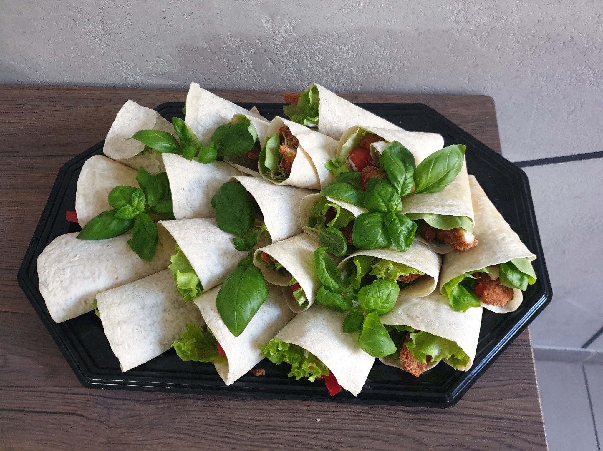 Tortille z kurczakiem, sałatą i bazylią ułożone na czarnej tacy, widoczne z bliska, z elementami czerwonej papryki.