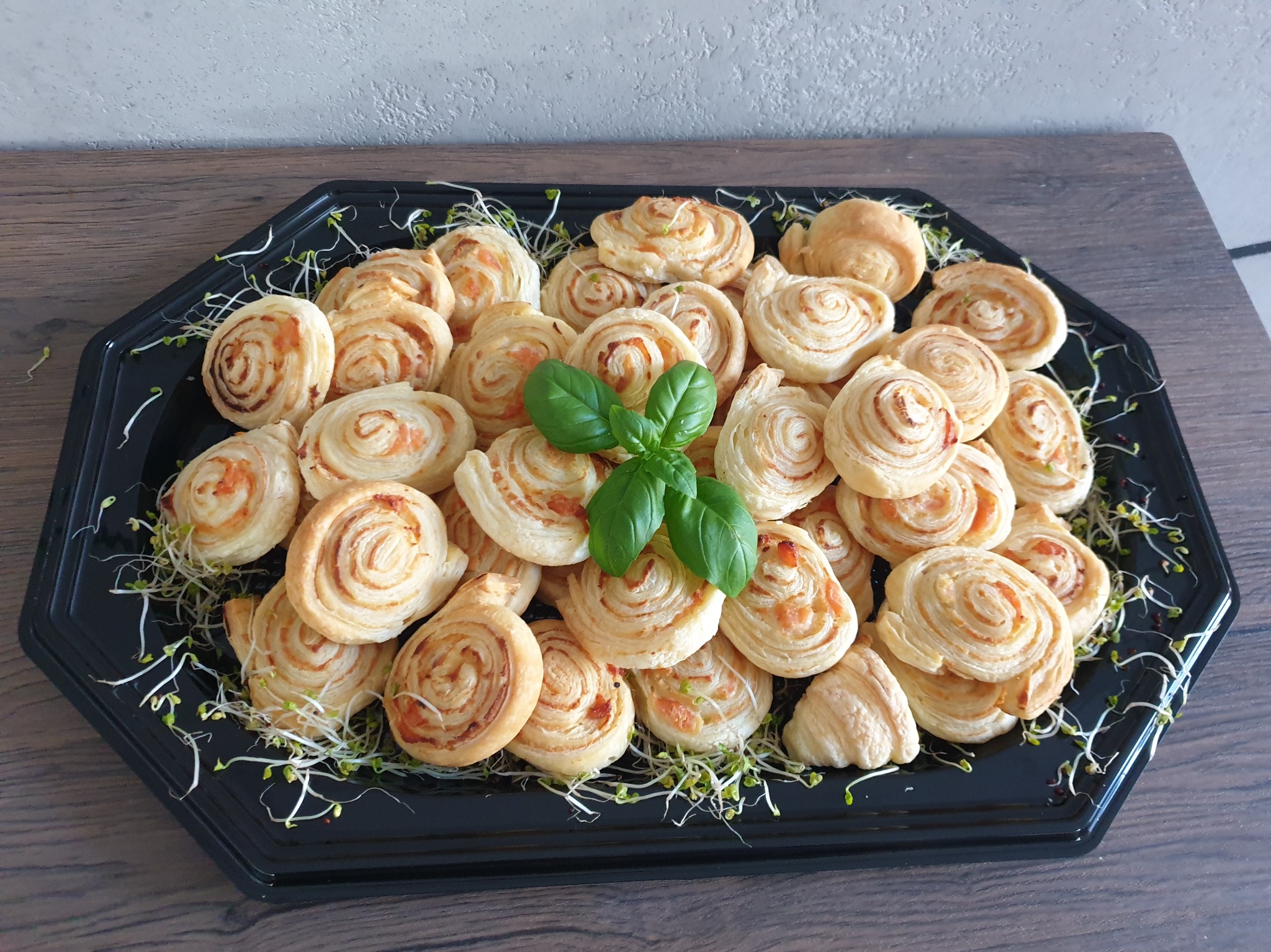 Czarny, ośmiokątny talerz z roladkami z ciasta francuskiego z farszem, udekorowany młodymi pędami i listkami bazylii.