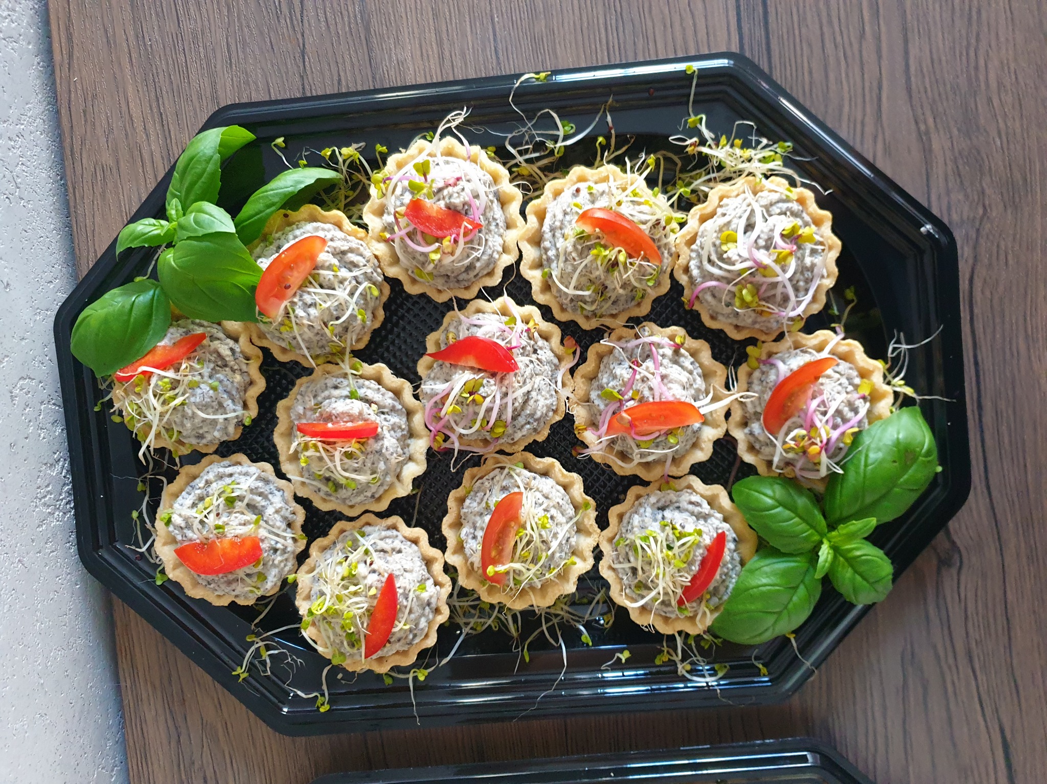 Tarty z szarą pastą, kiełkami i kawałkiem pomidora, ułożone na czarnej tacy z listkami bazylii.