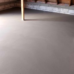 PRZEMYSŁAW PIOTRAK Alfa-Mix - Świeżo wylana, gładka posadzka jastrychowa w surowym pomieszczeniu poddasza z widoczną drewnianą konstrukcją dachu i fragmentami muru z bloczków betonowych.