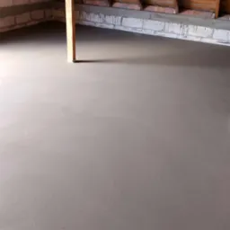 Świeżo wylana, gładka posadzka jastrychowa w surowym pomieszczeniu poddasza z widoczną drewnianą konstrukcją dachu i fragmentami muru z bloczków betonowych.