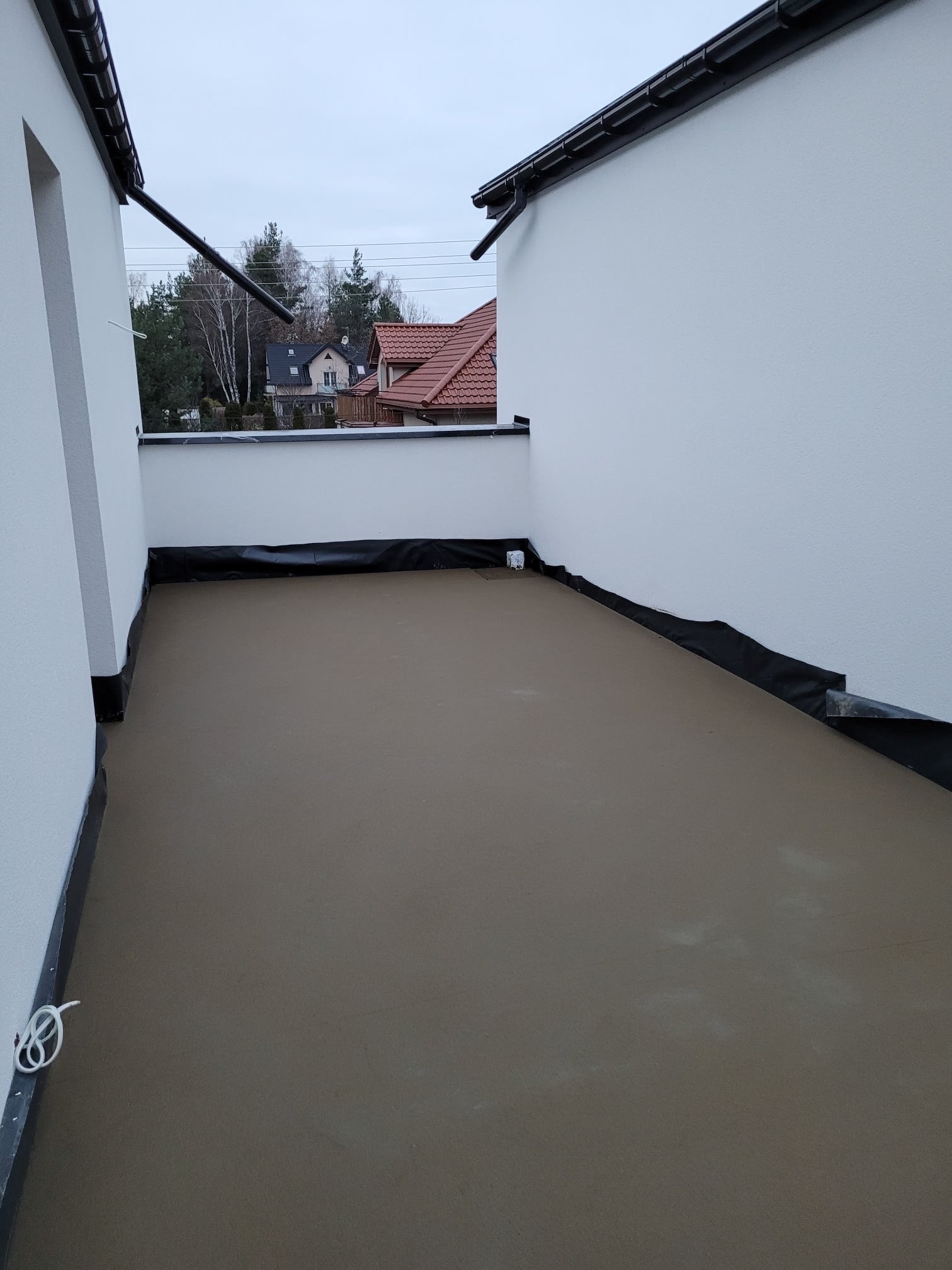 Świeżo wylana, gładka, szara powierzchnia na tarasie z widocznymi elementami izolacji przy ścianach i odpływem. W tle zabudowa jednorodzinna.