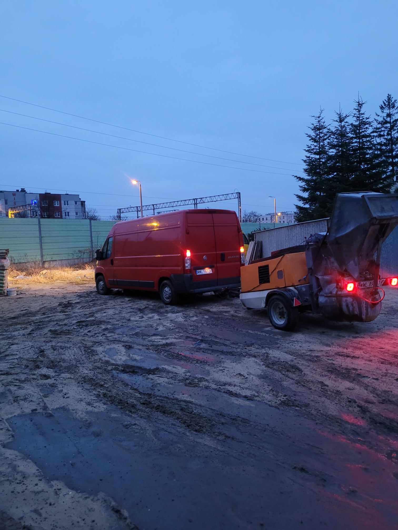 Czerwony van z przyczepioną maszyną do wylewek na placu budowy, widoczne błoto i zapadający zmierzch.