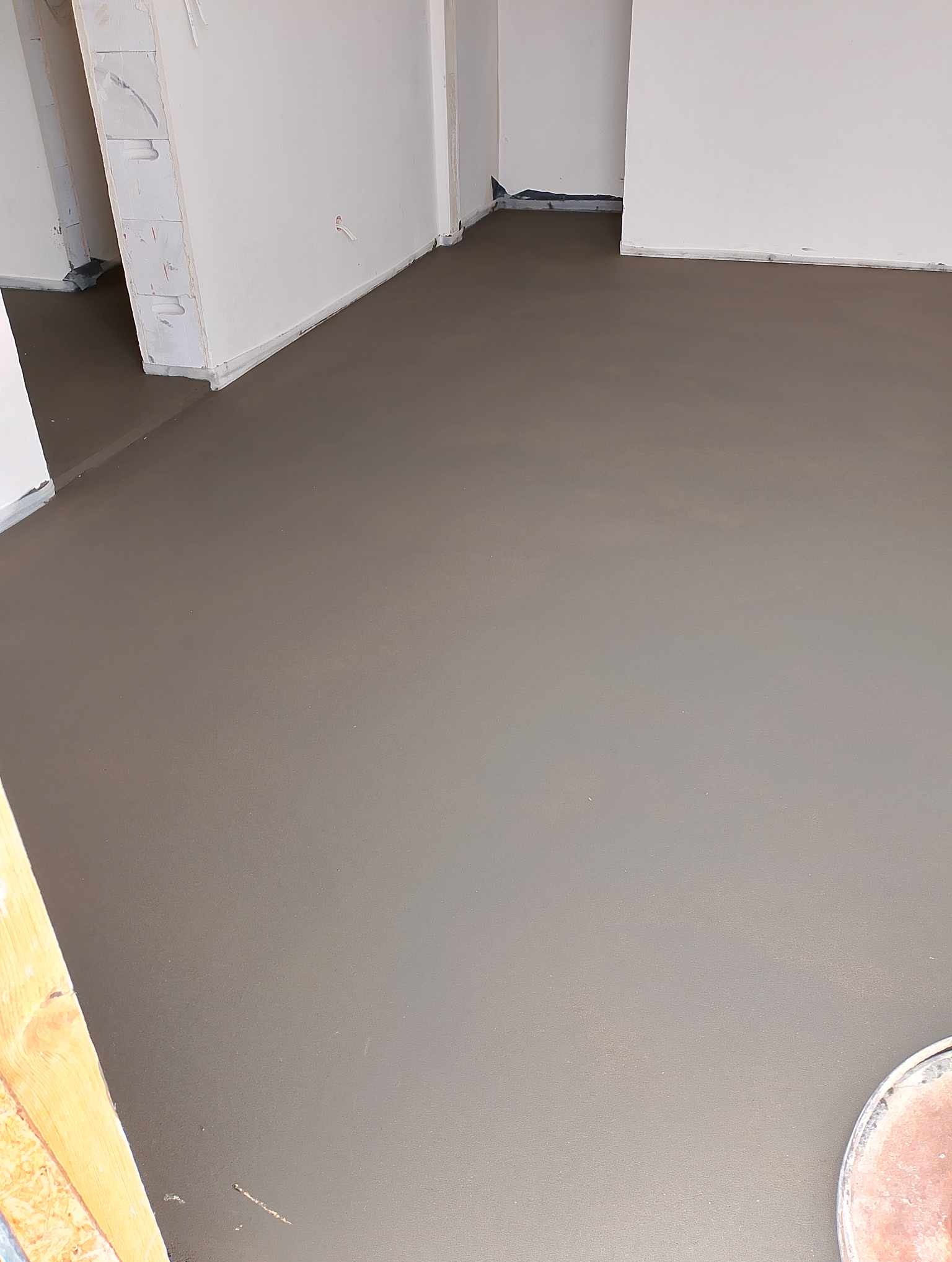 Świeżo wylana, gładka, szara posadzka w jasnym pomieszczeniu z białymi ścianami. Widoczna listwa przypodłogowa i fragment wiadra w prawym dolnym rogu.