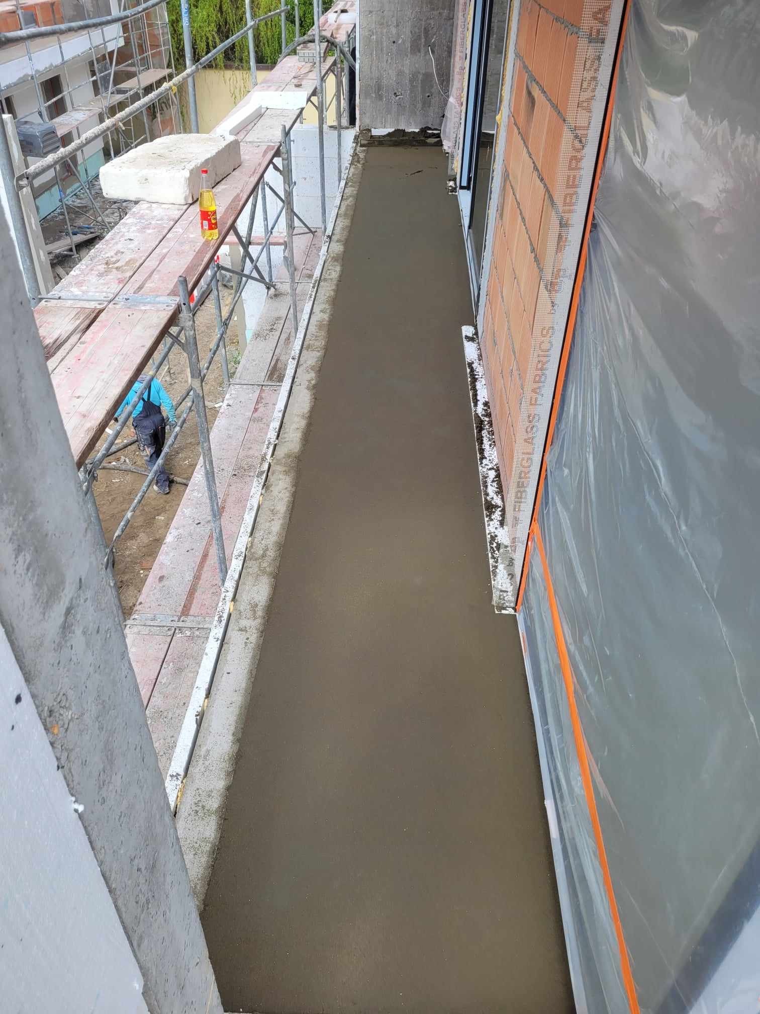 Świeżo wylana, gładka posadzka na balkonie, zabezpieczona folią budowlaną, widok z góry na rusztowanie z butelką oleju na krawędzi.