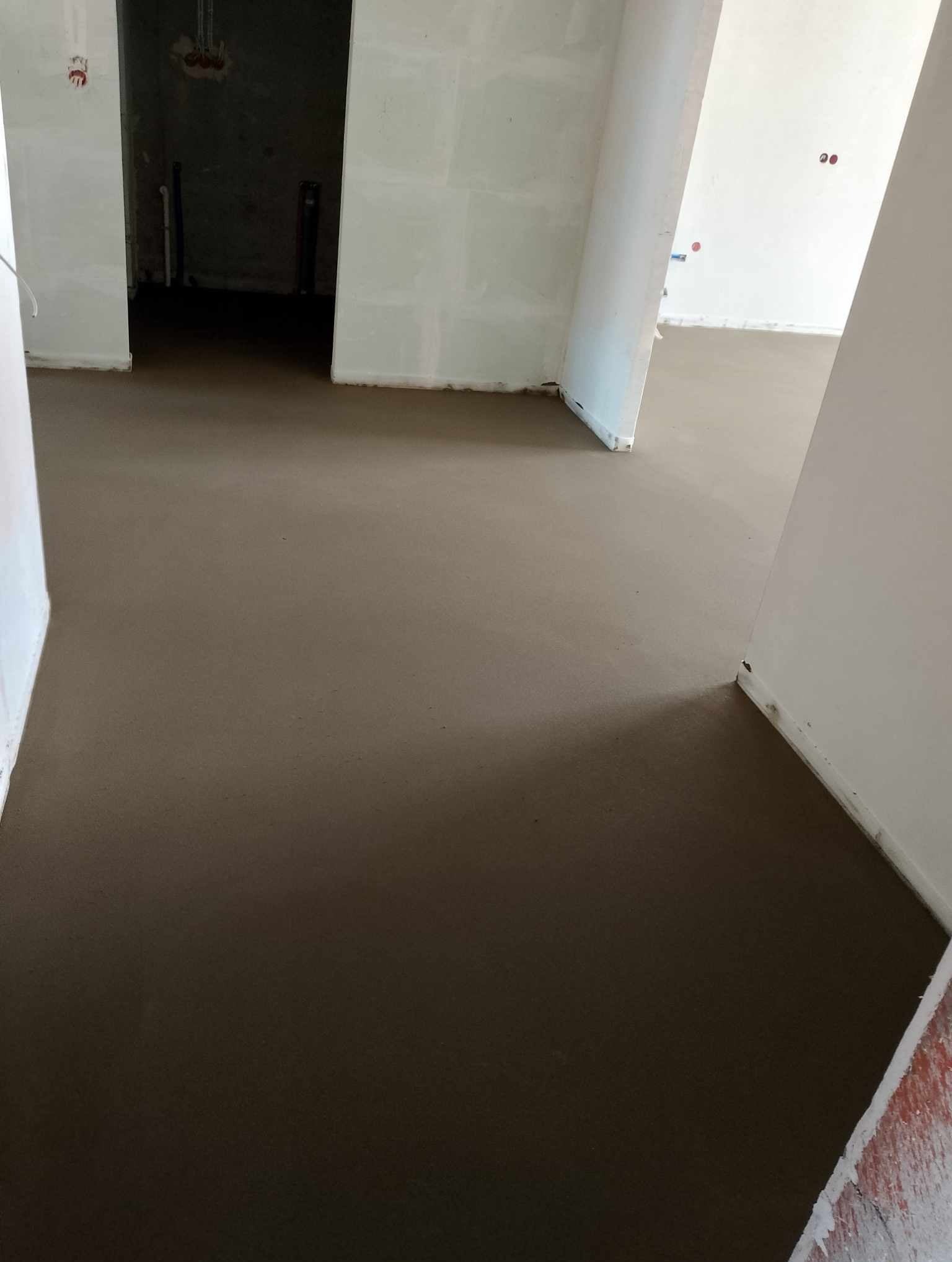 Świeżo wylana, gładka i równa posadzka w jasnym wnętrzu z widocznymi instalacjami elektrycznymi w tle, w trakcie prac wykończeniowych.