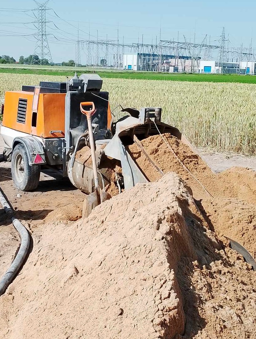 Pomarańczowo-szara maszyna do wylewek z dwiema łopatami wbitymi w pryzmę piasku na polu, w tle słupy wysokiego napięcia i budynek stacji transformatorowej.