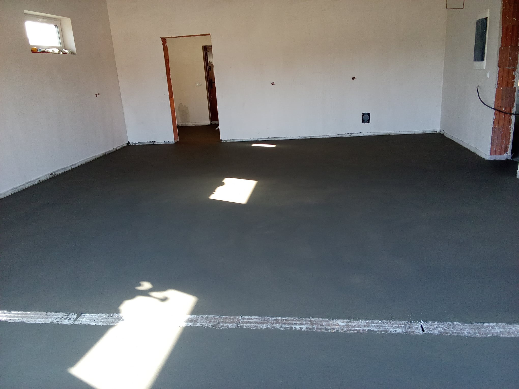 Świeżo wylana, gładka posadzka w surowym stanie, z widocznym paskiem dylatacyjnym i padającym na nią światłem słonecznym, w tle surowe ściany budynku z otworami okiennymi i drzwiowymi.