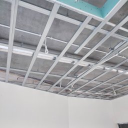 Gor-Bud - Stelaż sufitu podwieszanego z widocznymi przewodami i elementami instalacji, surowy betonowy strop, ściany przygotowane do malowania z widocznymi puszkami elektrycznymi.