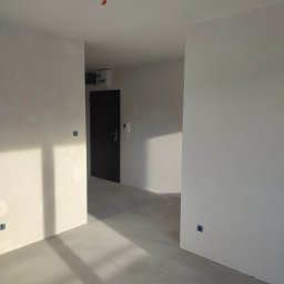 Gor-Bud - Wnętrze w stanie deweloperskim z gładkimi, pomalowanymi na biało ścianami, surową betonową podłogą, widocznymi puszkami elektrycznymi i wystającymi kablami z sufitu, czarne drzwi wejściowe w głębi.