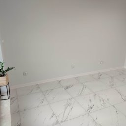 Gor-Bud - Puste, jasne pomieszczenie z nową podłogą z płytek imitujących marmur, białe ściany z widocznymi gniazdkami elektrycznymi i rośliną doniczkową w drewnianej osłonce na metalowym stojaku przy oknie.