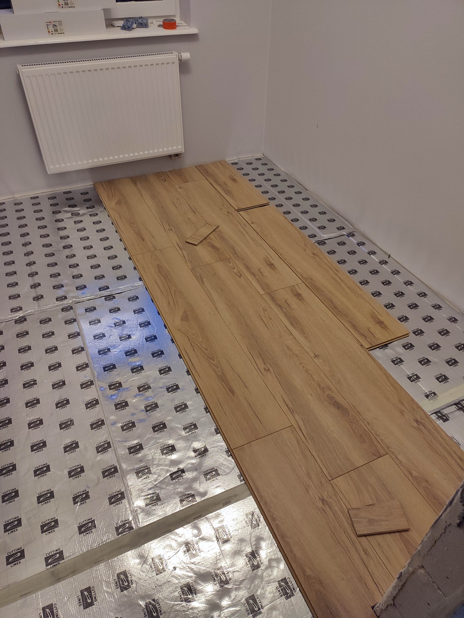 Proces układania paneli podłogowych na folii izolacyjnej z widocznym fragmentem ściany z grzejnikiem.