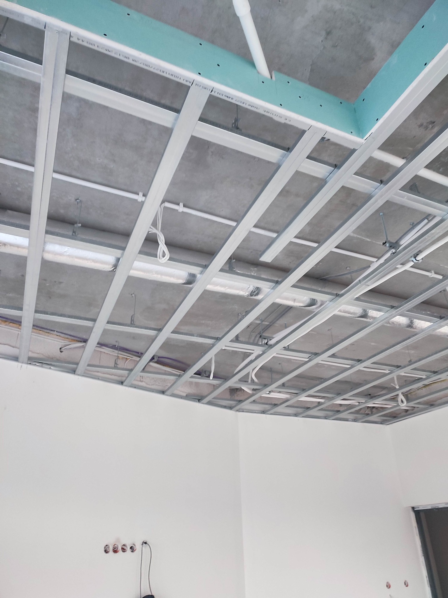 Stelaż sufitu podwieszanego z widocznymi przewodami i elementami instalacji, surowy betonowy strop, ściany przygotowane do malowania z widocznymi puszkami elektrycznymi.